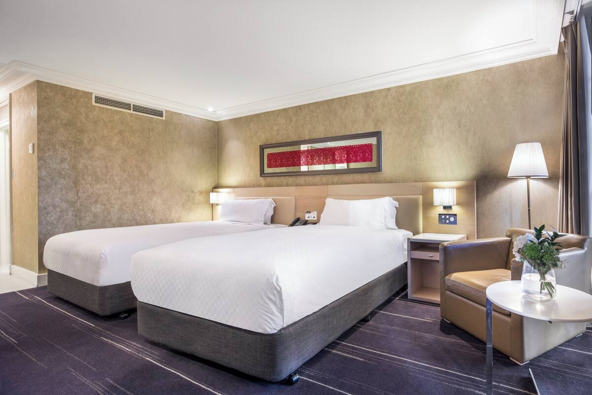 A hotel room with two beds, a small table with a vase, a chair, and a nightstand with a lamp.
