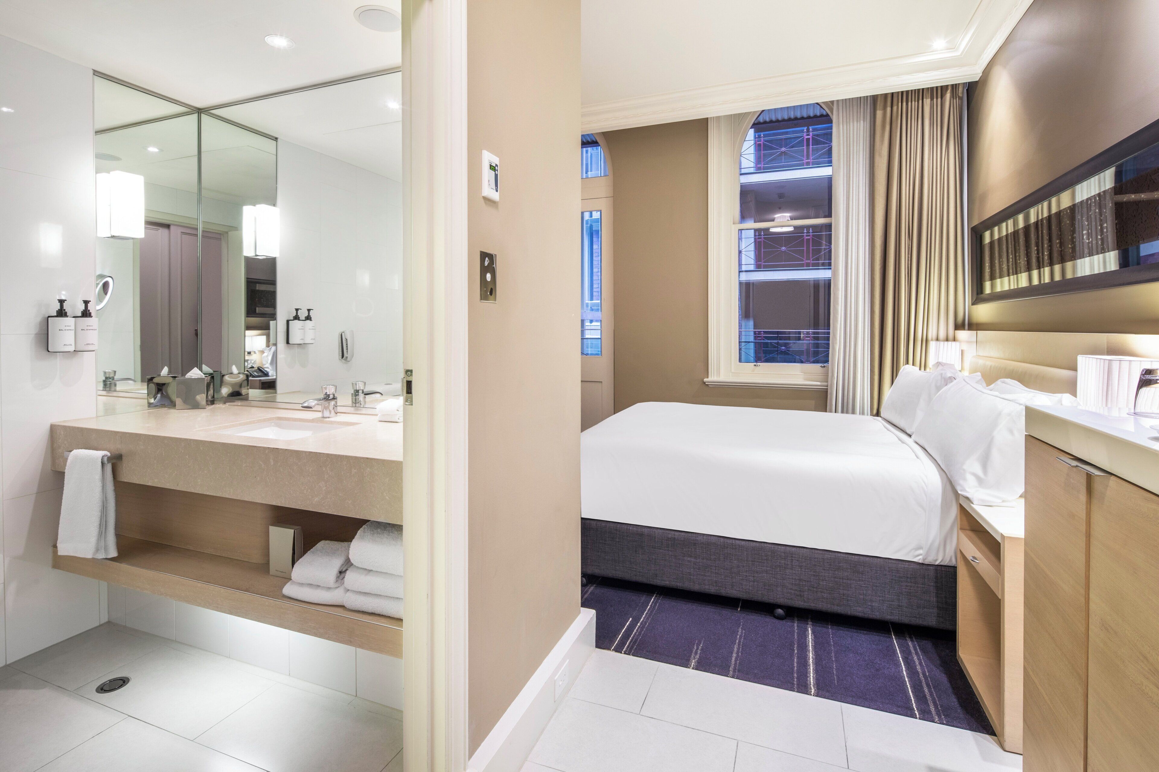 A modern hotel room with a large bed, a separate bathroom with a large mirror, and a view of the cityscape.