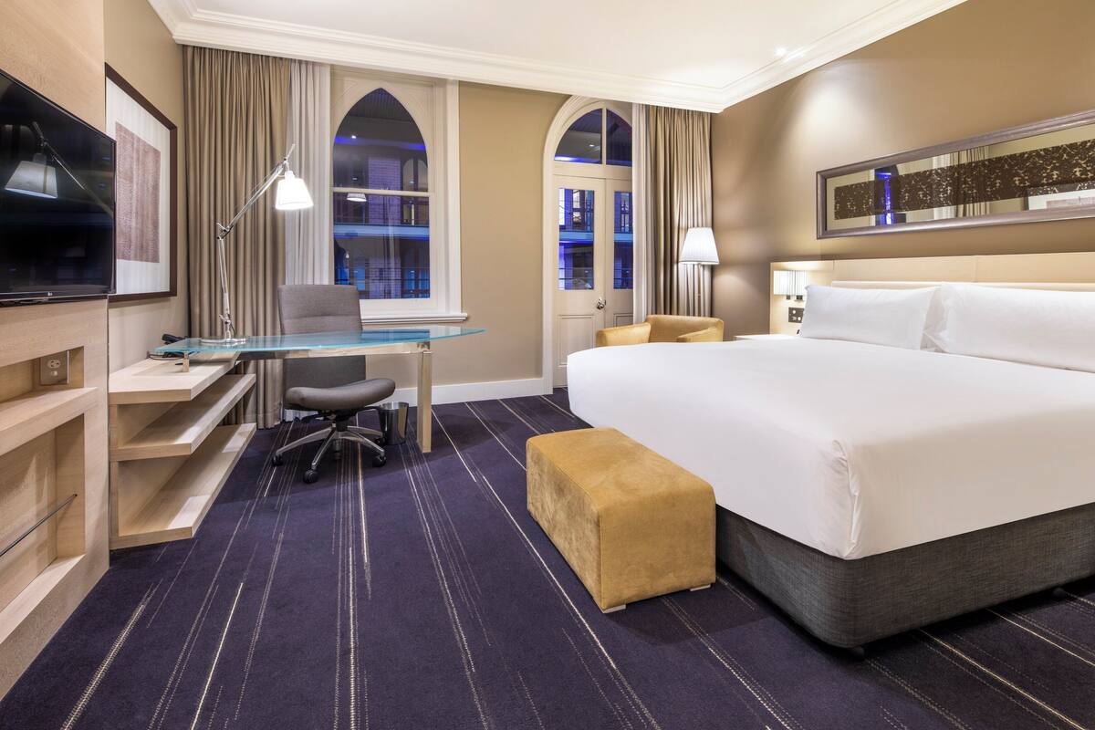 A hotel room with a large bed, a desk with a chair, a television, and a window with curtains.