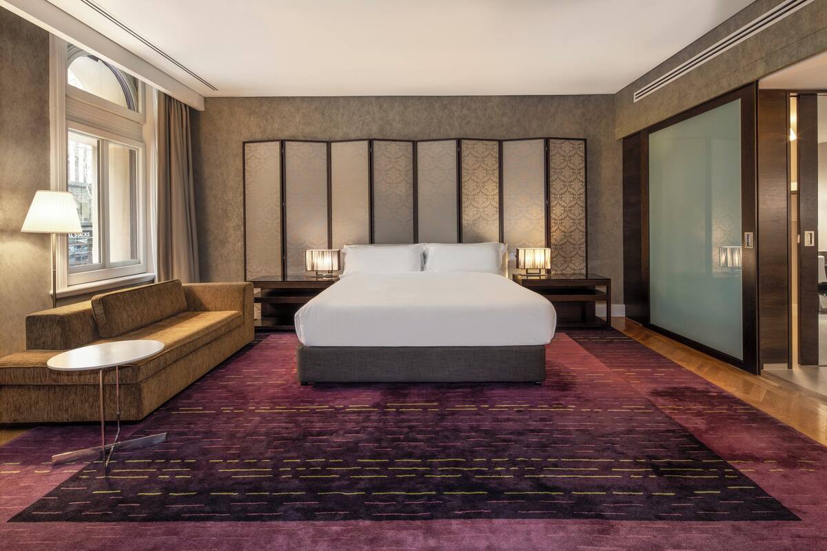 A modern hotel room with a large bed, a sofa, a small table, and a large window.