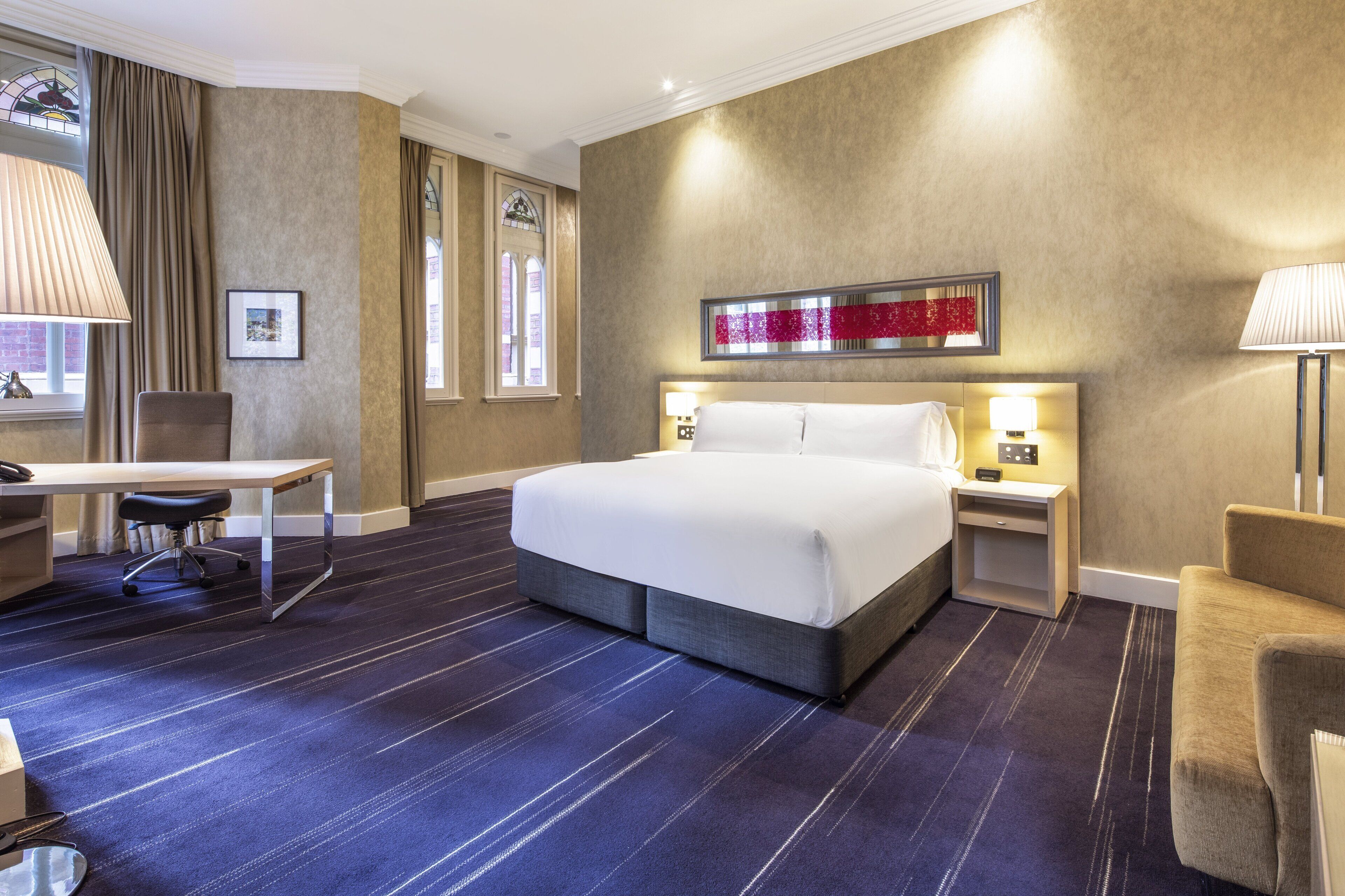A hotel room with a large bed, a desk, a chair, and a nightstand with a lamp.
