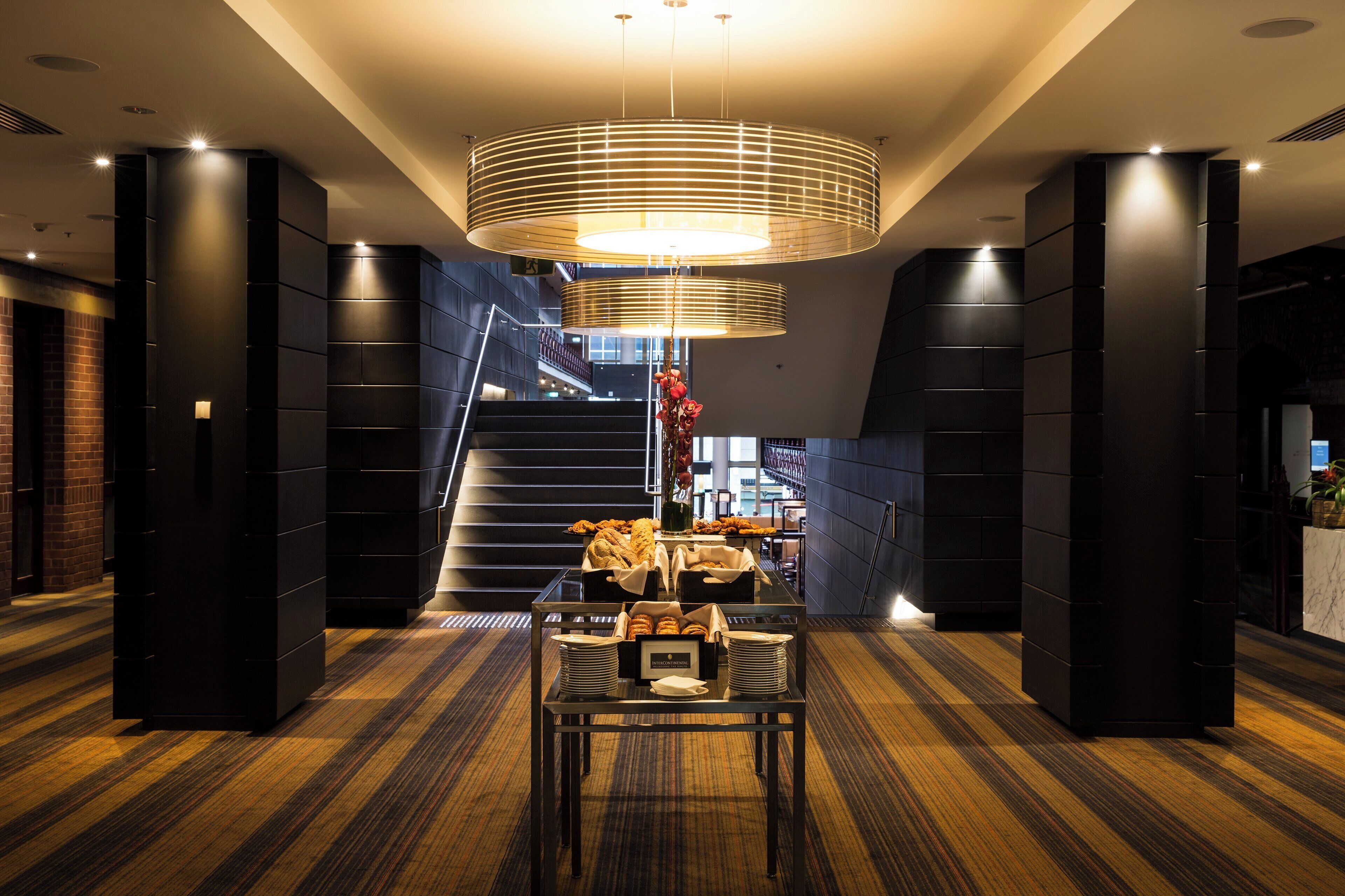A modern hotel lobby with a central staircase, a dining area with bread and pastries, and a large circular light fixture.