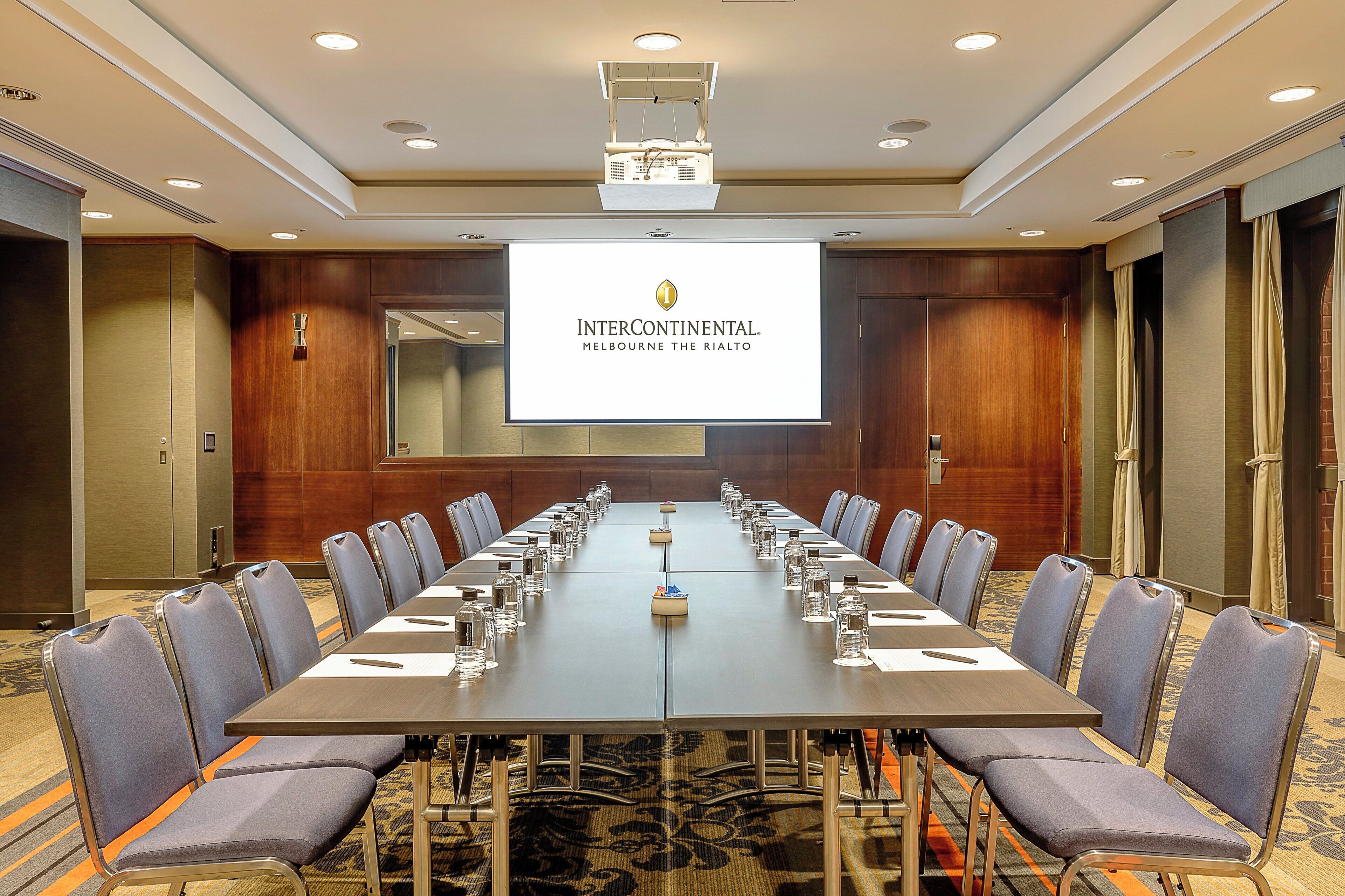 Un salón de conferencias con una larga mesa, sillas, una pantalla de proyector y un logotipo en la pared.
