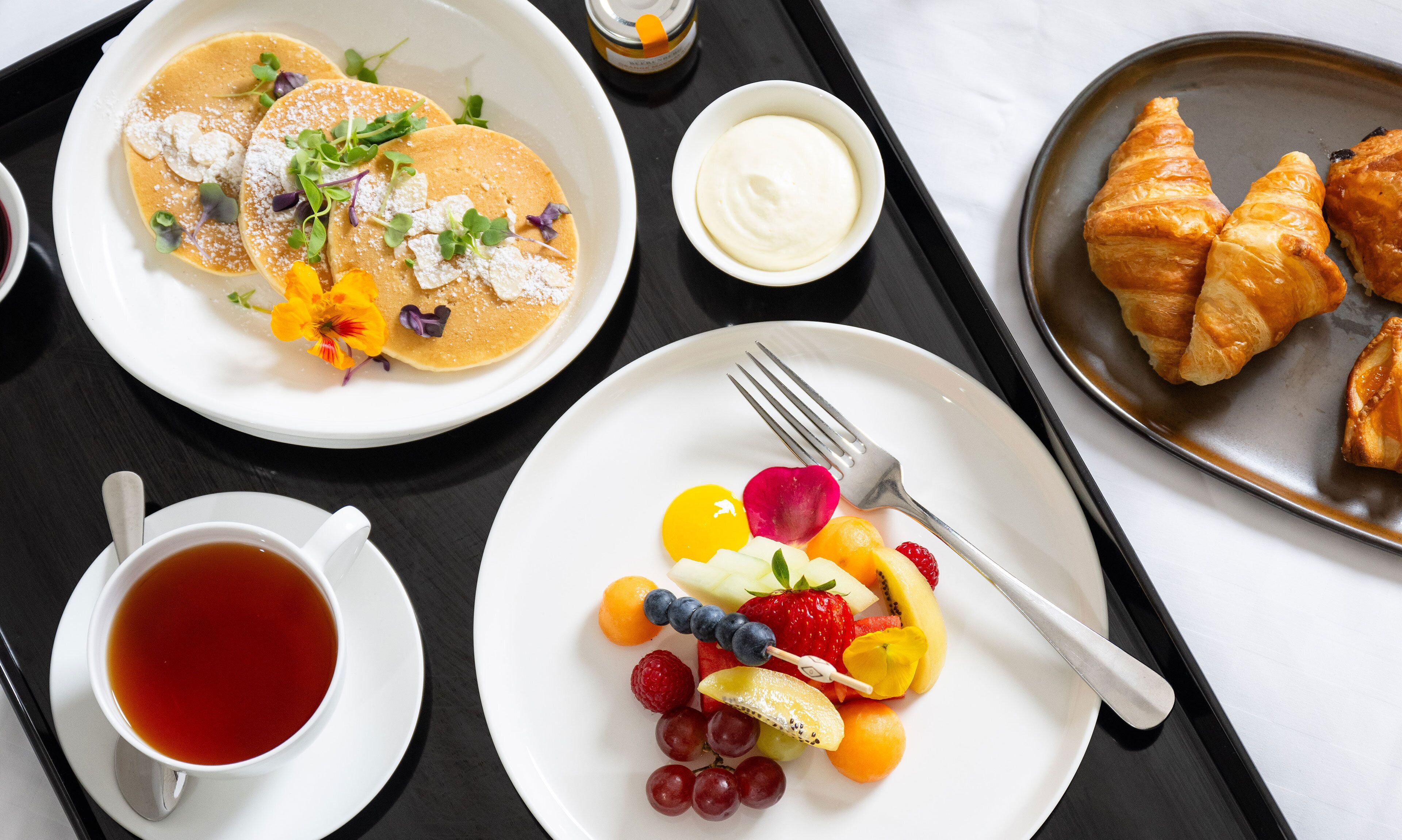 Bandeja de desayuno con panqueques, cruasanes, fruta y una taza de té.