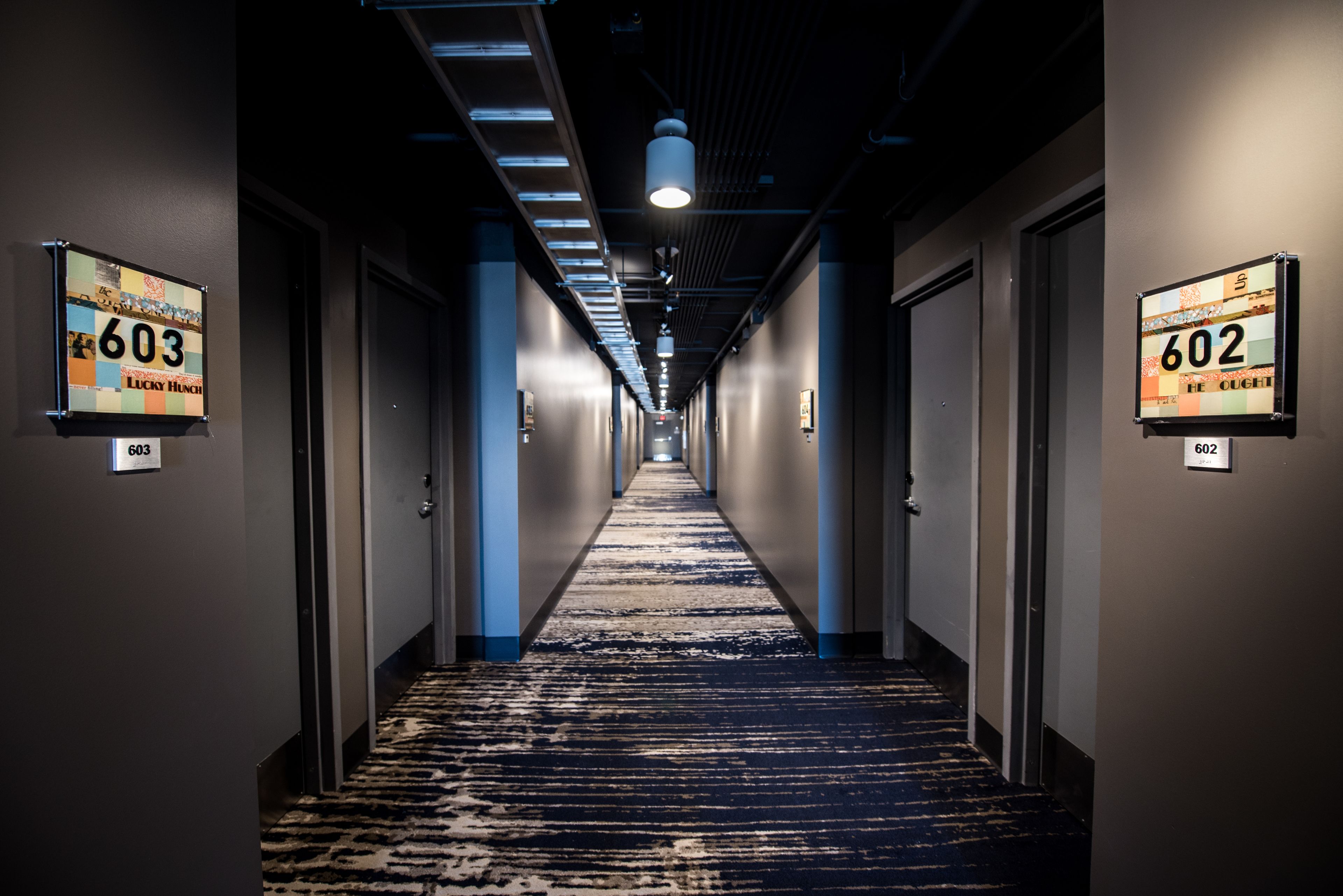 Een hotelgang met vloerbedekking, deuren met de aanduidingen 603 en 602, en verlichting in het plafond.