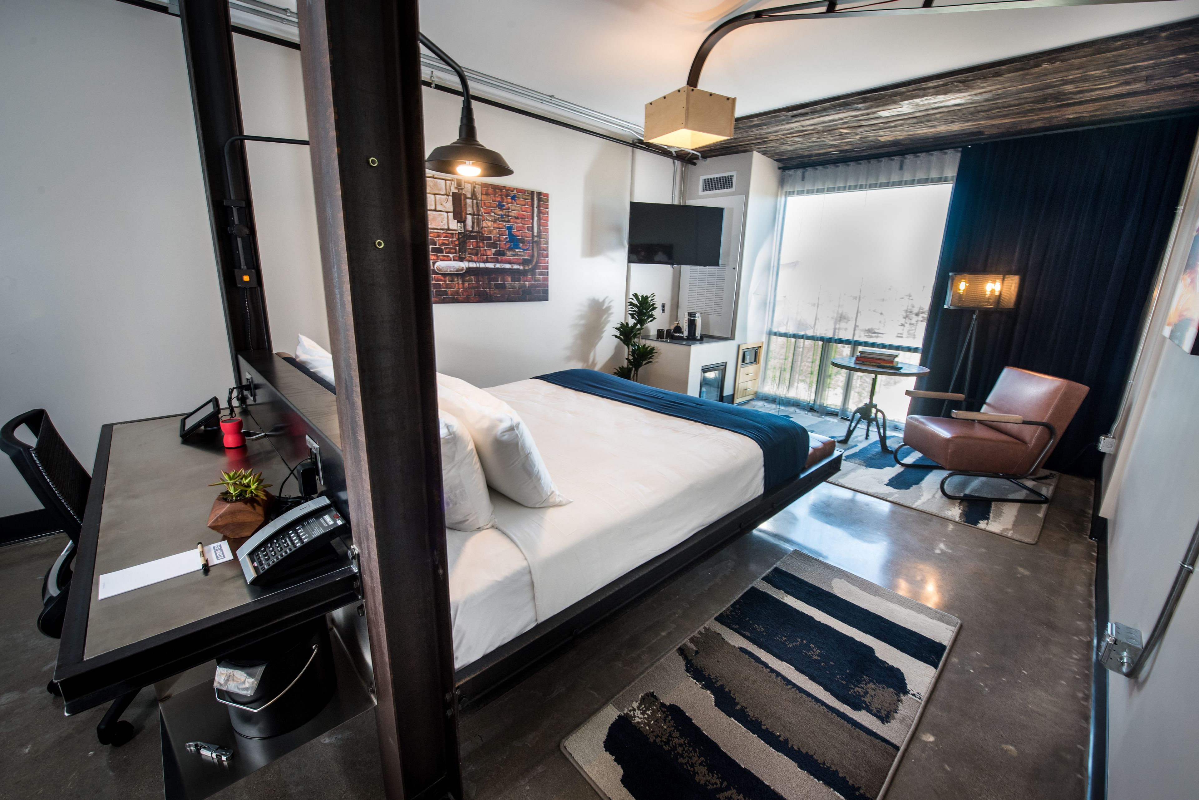 Una habitació d'hotel moderna amb un llit, escriptori, cadira i una petita zona de balcó.