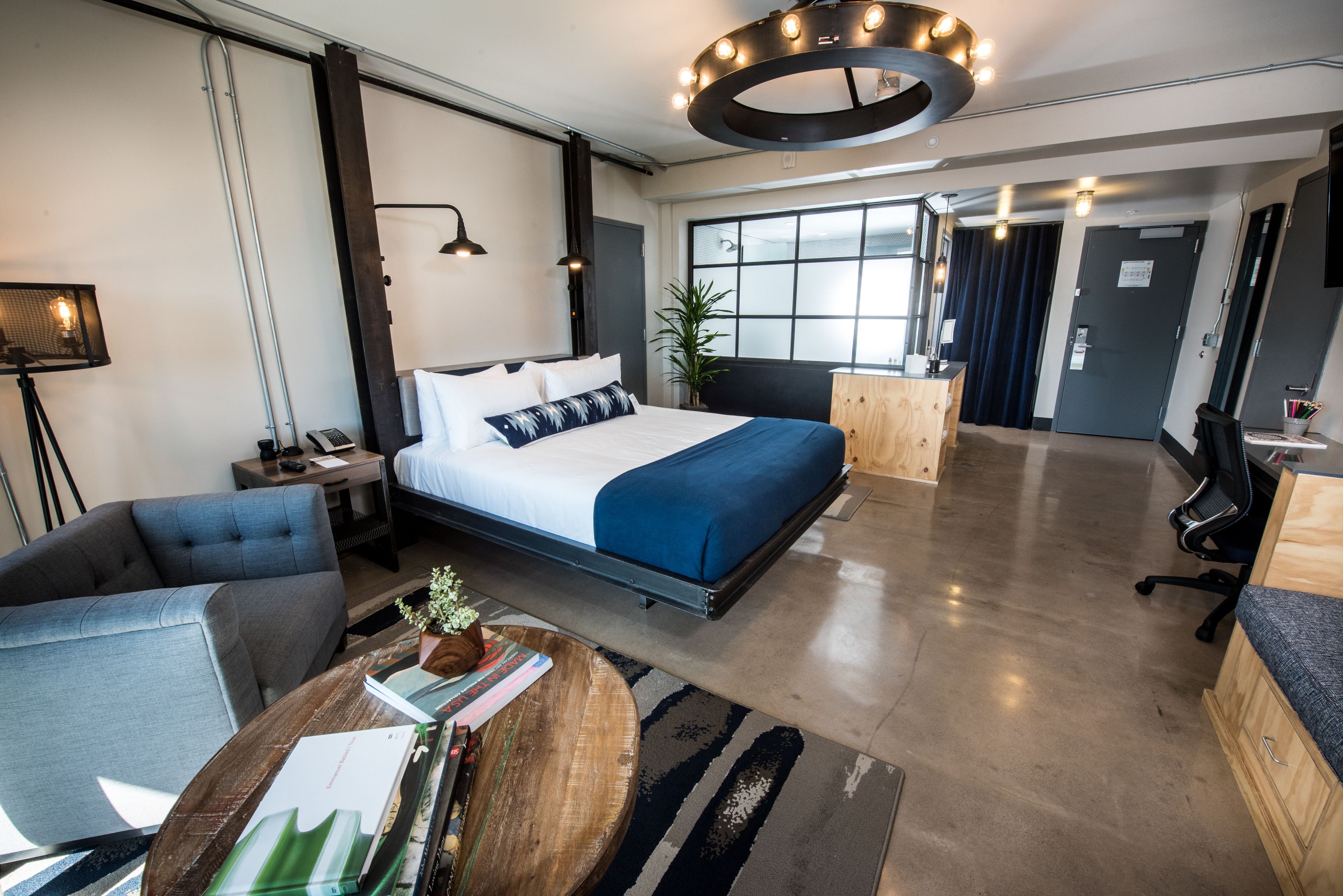 A modern hotel room with a large bed, a desk, a chair, and a round coffee table.