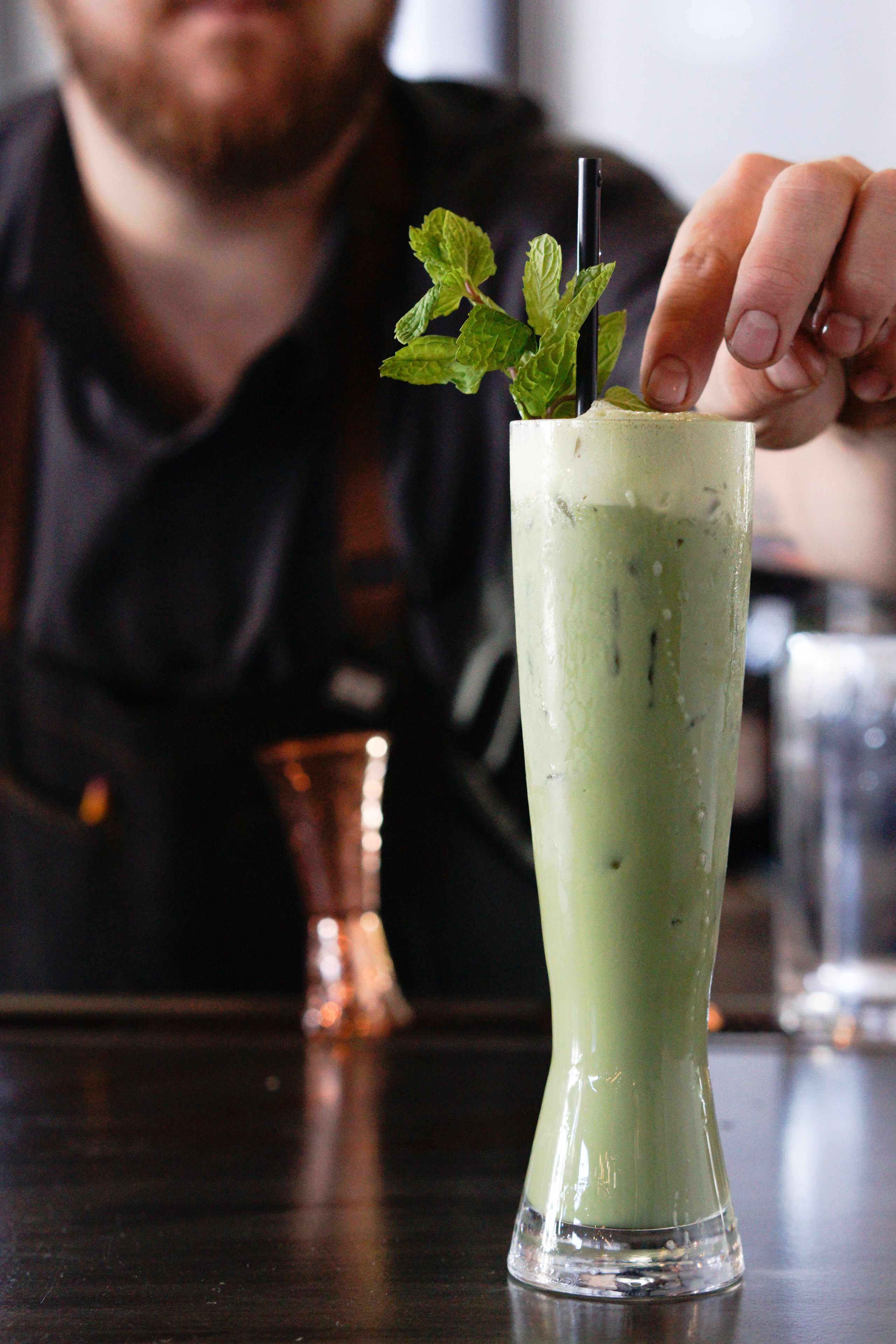 Een hoog glas met een groen drankje, versierd met muntblaadjes en een rietje, op een donker oppervlak.