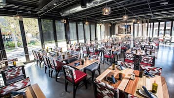 Un restaurante espacioso con varias mesas y sillas, ventanas amplias y un techo moderno de estilo industrial.