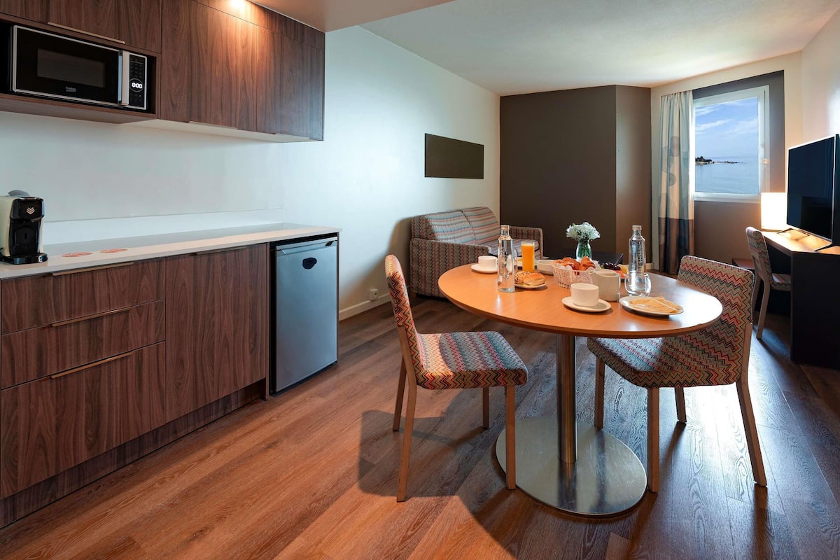 A modern kitchen with a microwave, coffee maker, and a small refrigerator. There is a dining area with a round table and patterned chairs. A sofa and a TV are also visible.