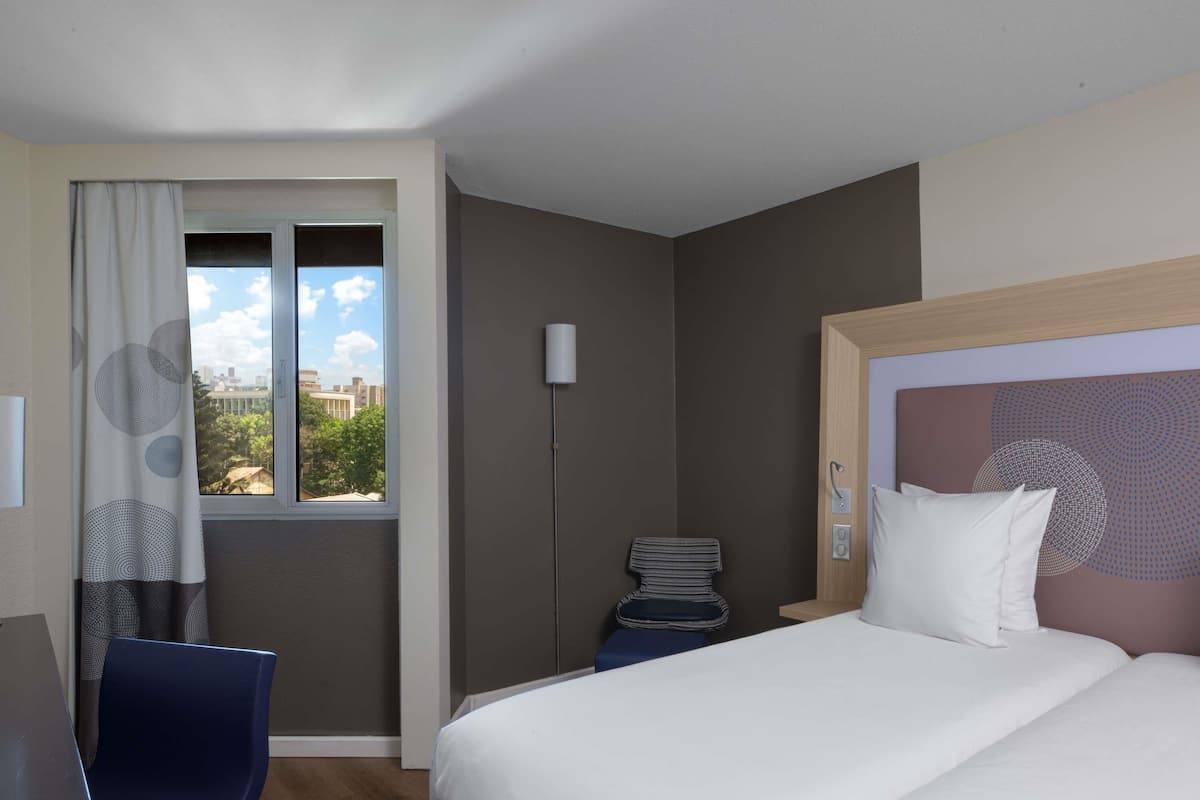 A hotel room with a bed, a desk, a chair, a standing lamp, and a view of buildings through the window.