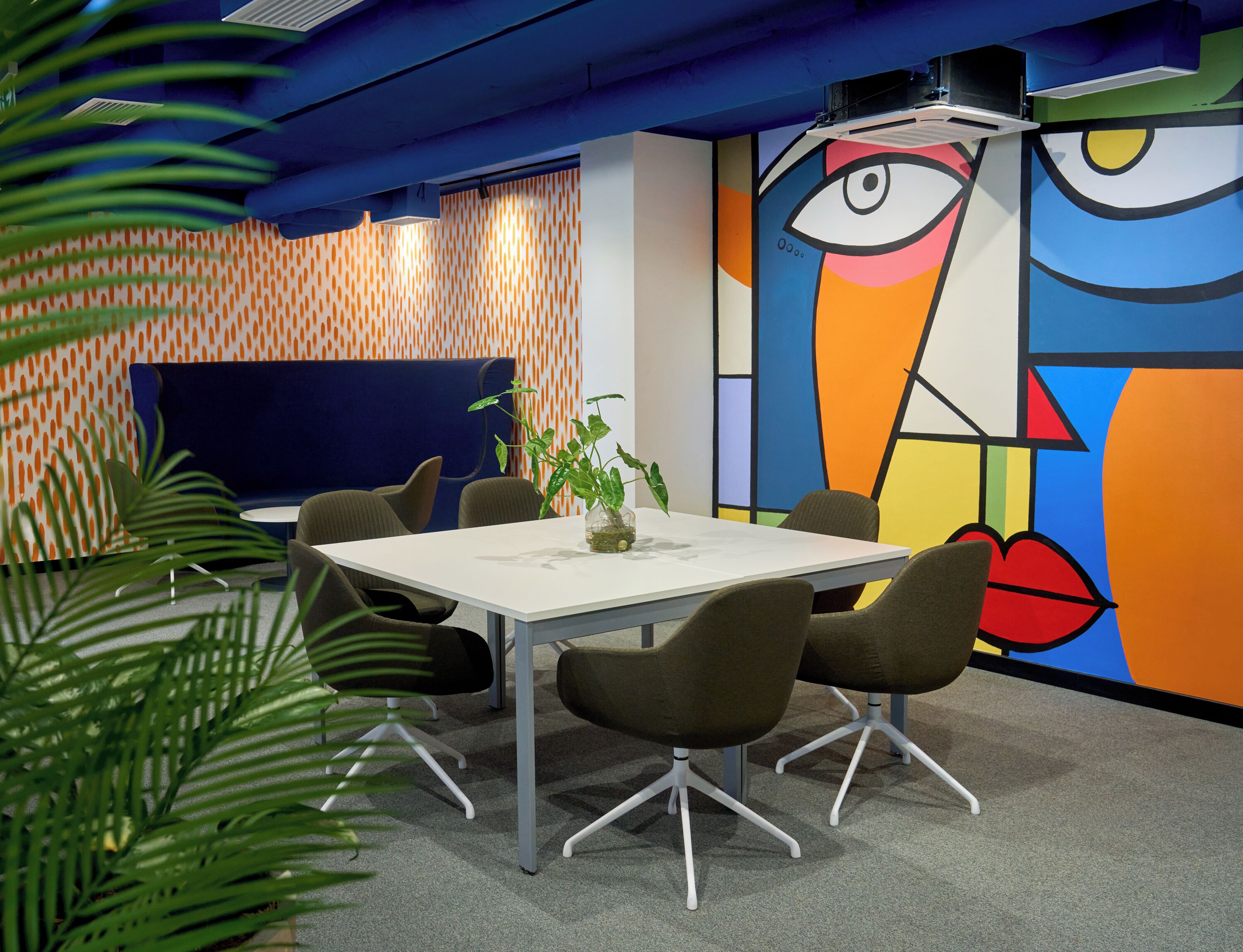 A modern meeting room with a white table, brown chairs, a potted plant, and a colorful abstract mural on the wall.