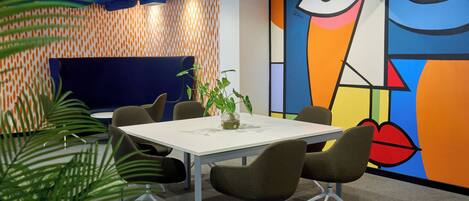 A modern meeting room with a white table, brown chairs, a potted plant, and a colorful abstract mural on the wall.