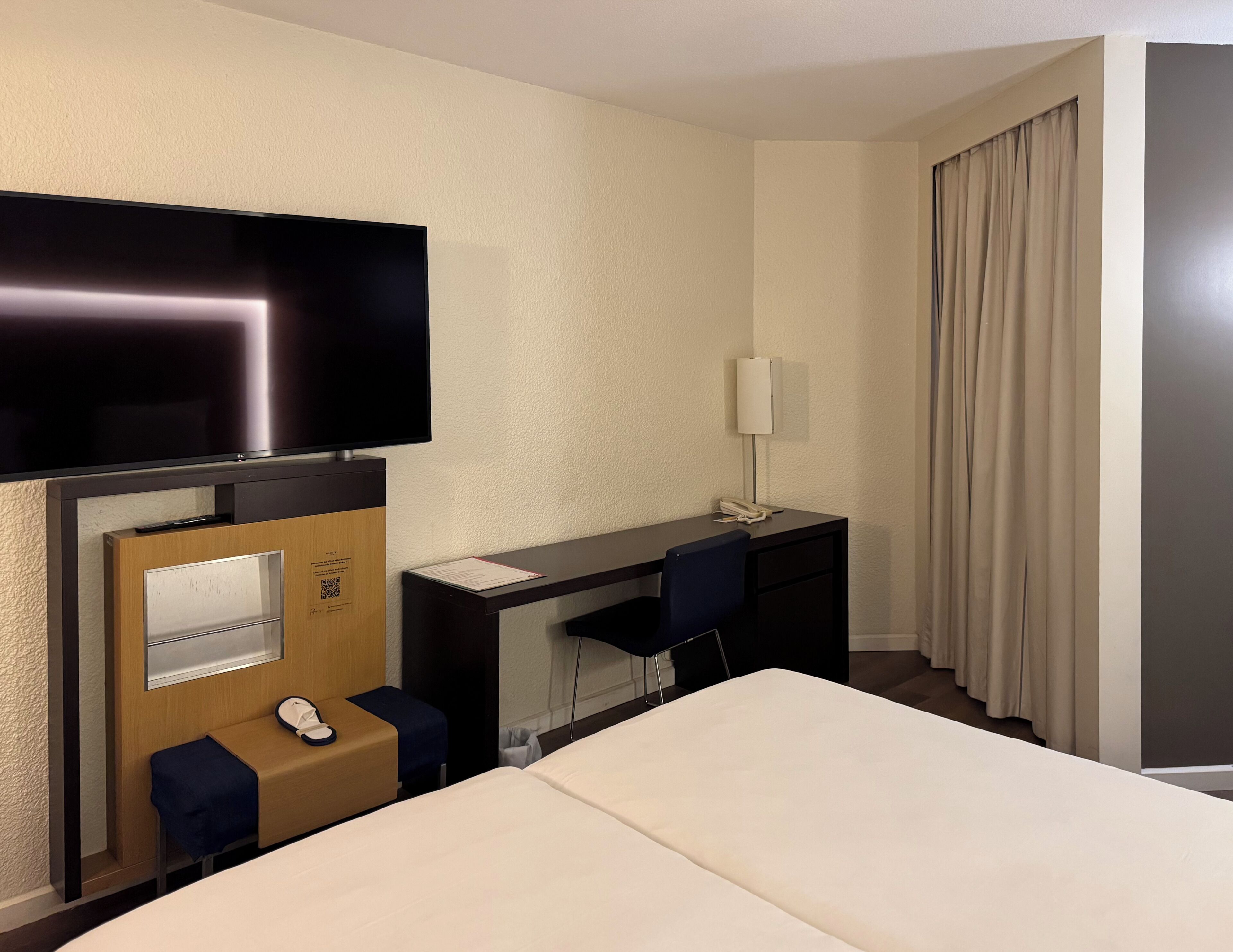 A hotel room with a bed, a desk with a chair, a television, a telephone, and a small table with a chair.