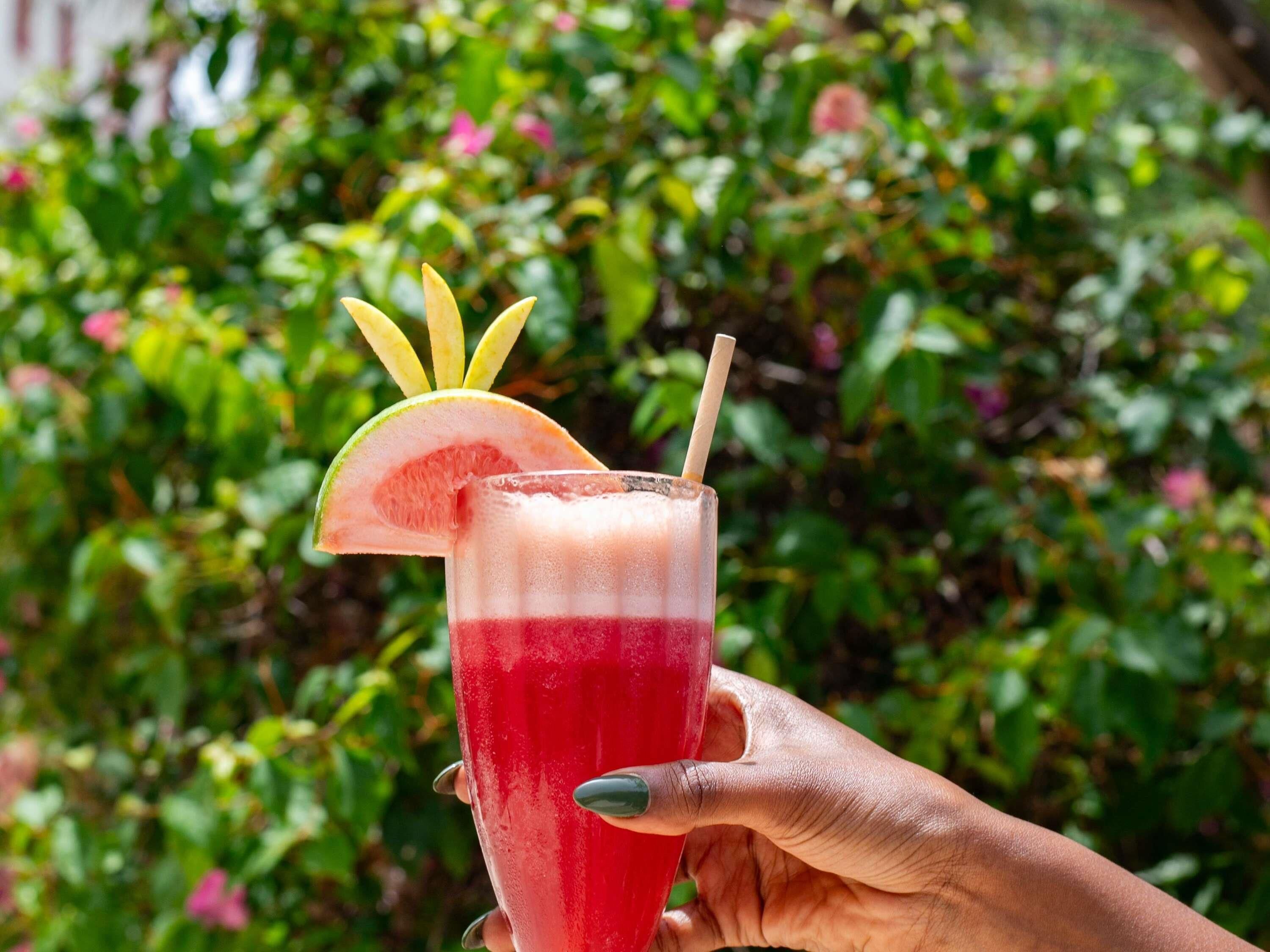 Een hand die een glas vasthoudt met een gelaagde drank, versierd met een stuk fruit en een rietje.