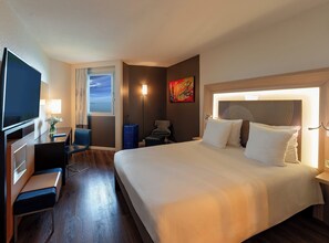 A hotel room with a bed, a desk with a chair, a TV, a window with curtains, a lamp, and a painting on the wall. - Novotel Dakar (Dakar)