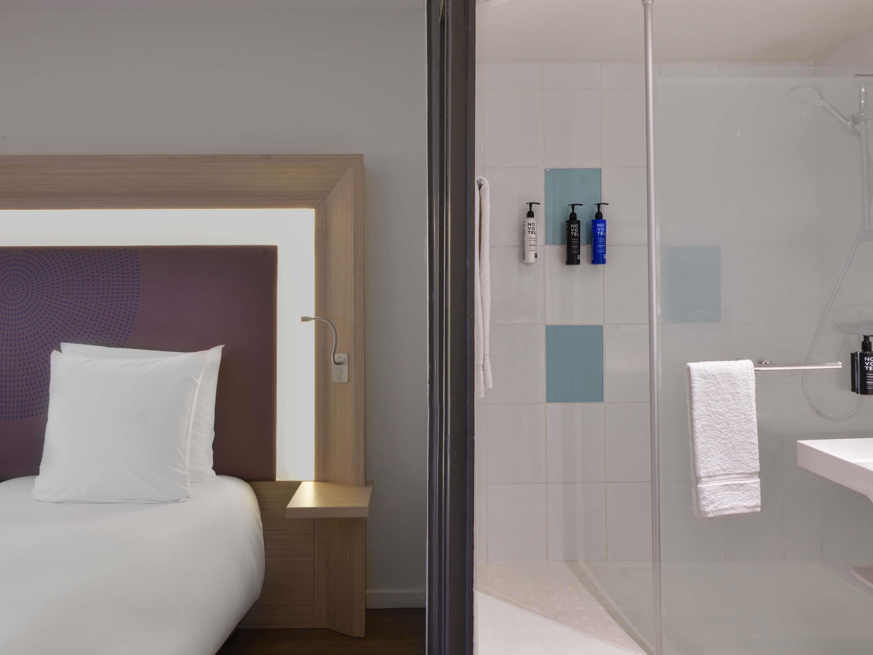 Una habitación de hotel moderna con una cama, una mesita de noche, cabina de ducha de vidrio y artículos de aseo en la pared.