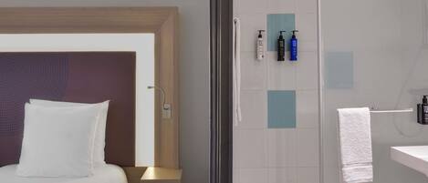 A modern hotel room with a bed, a nightstand, a glass shower enclosure, and toiletries on the wall.