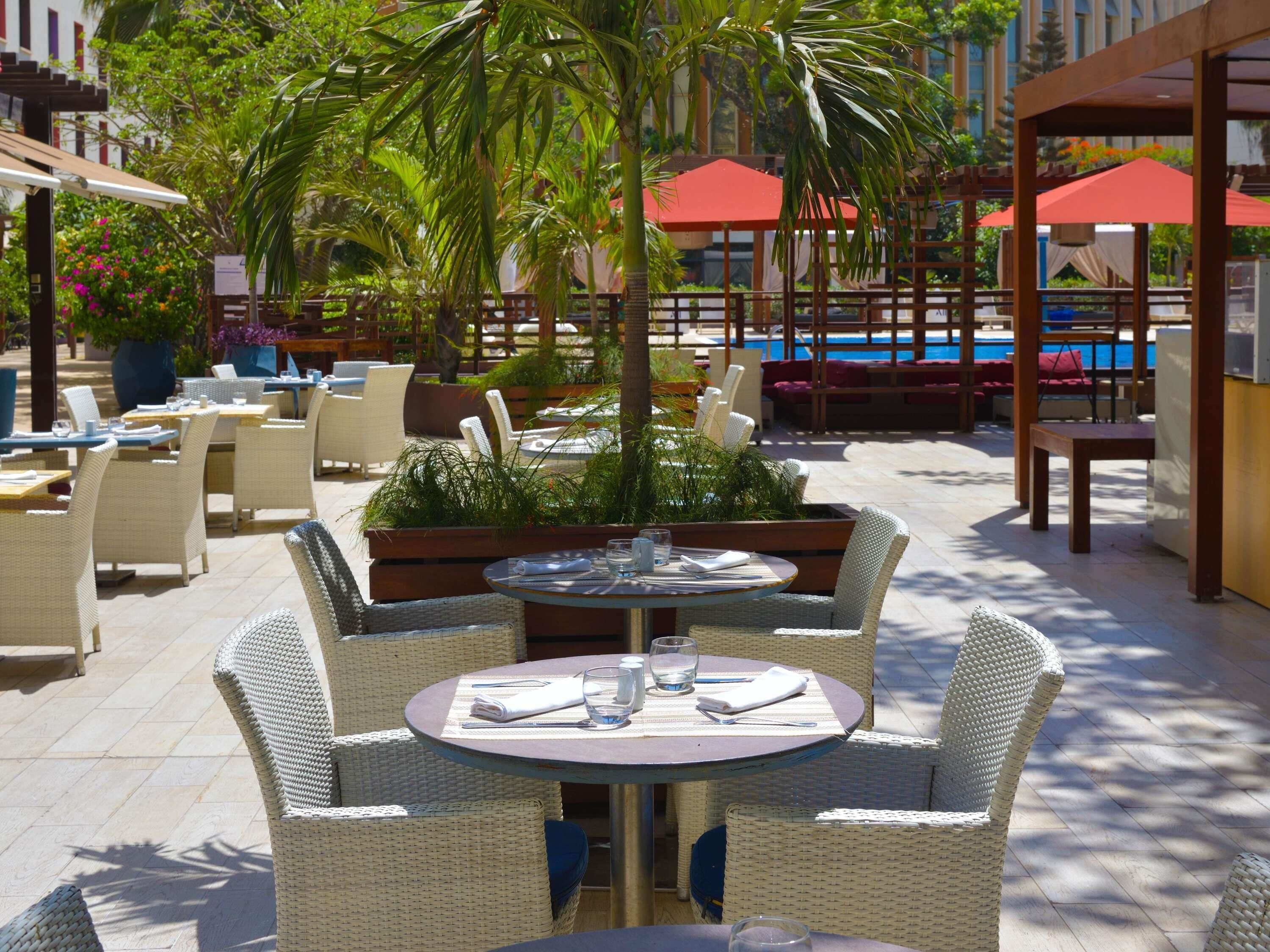 Zona pranzo all'aperto con mobili in vimini, piscina sullo sfondo e ombrelloni rossi.