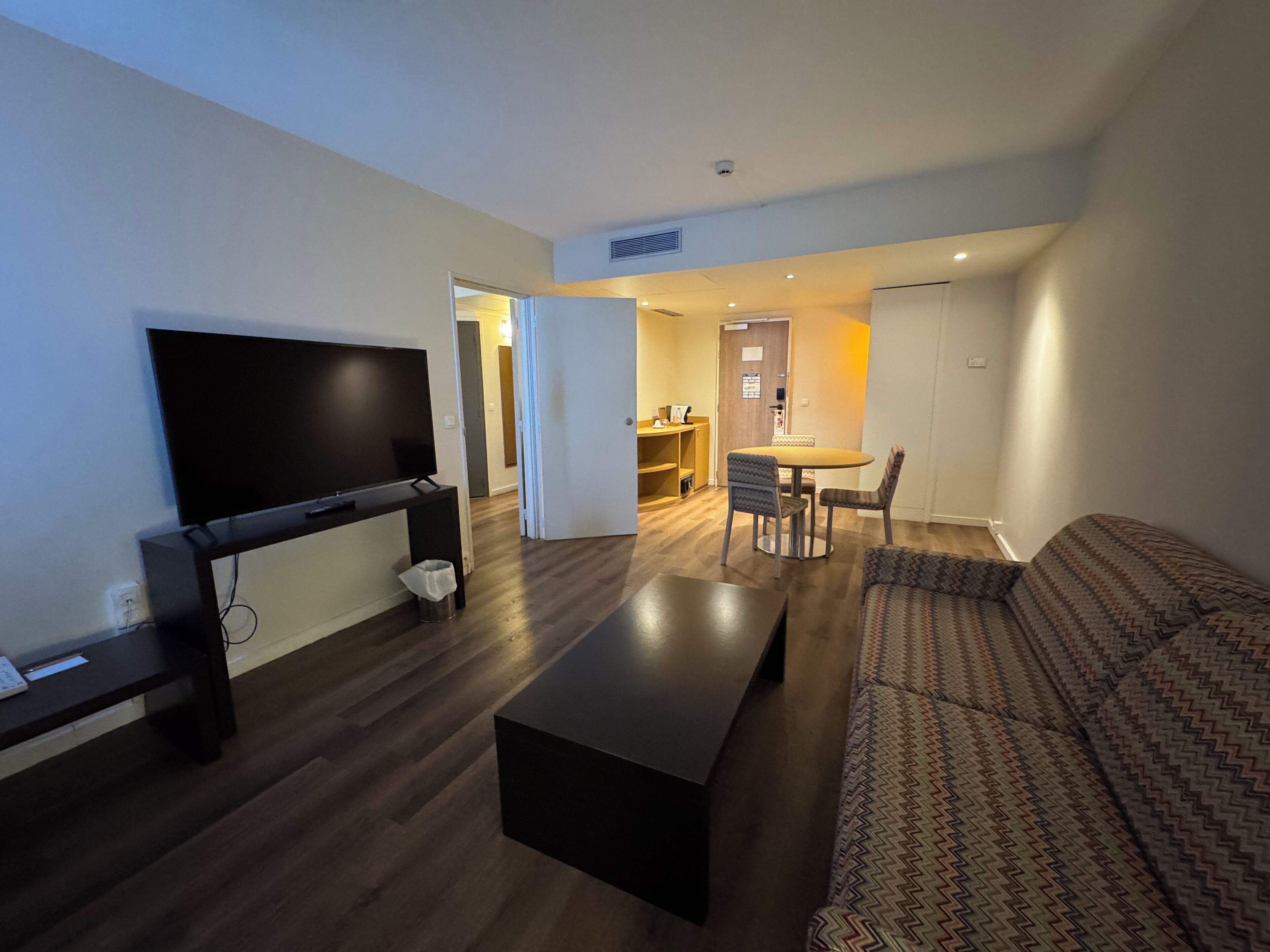 Ein modernes Wohnzimmer mit Flachbildfernseher, einem dunklen Couchtisch und einer gemusterten Couch. Im Hintergrund befindet sich eine Essecke mit Tisch und Stühlen.