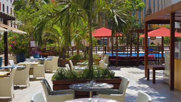 An outdoor dining area with wicker furniture, red umbrellas, and a pool in the background. - Novotel Dakar (Dakar)