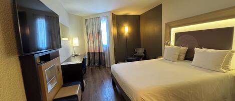 Une chambre d’hôtel avec un lit, un bureau, une chaise, une télévision et une fenêtre avec des rideaux.