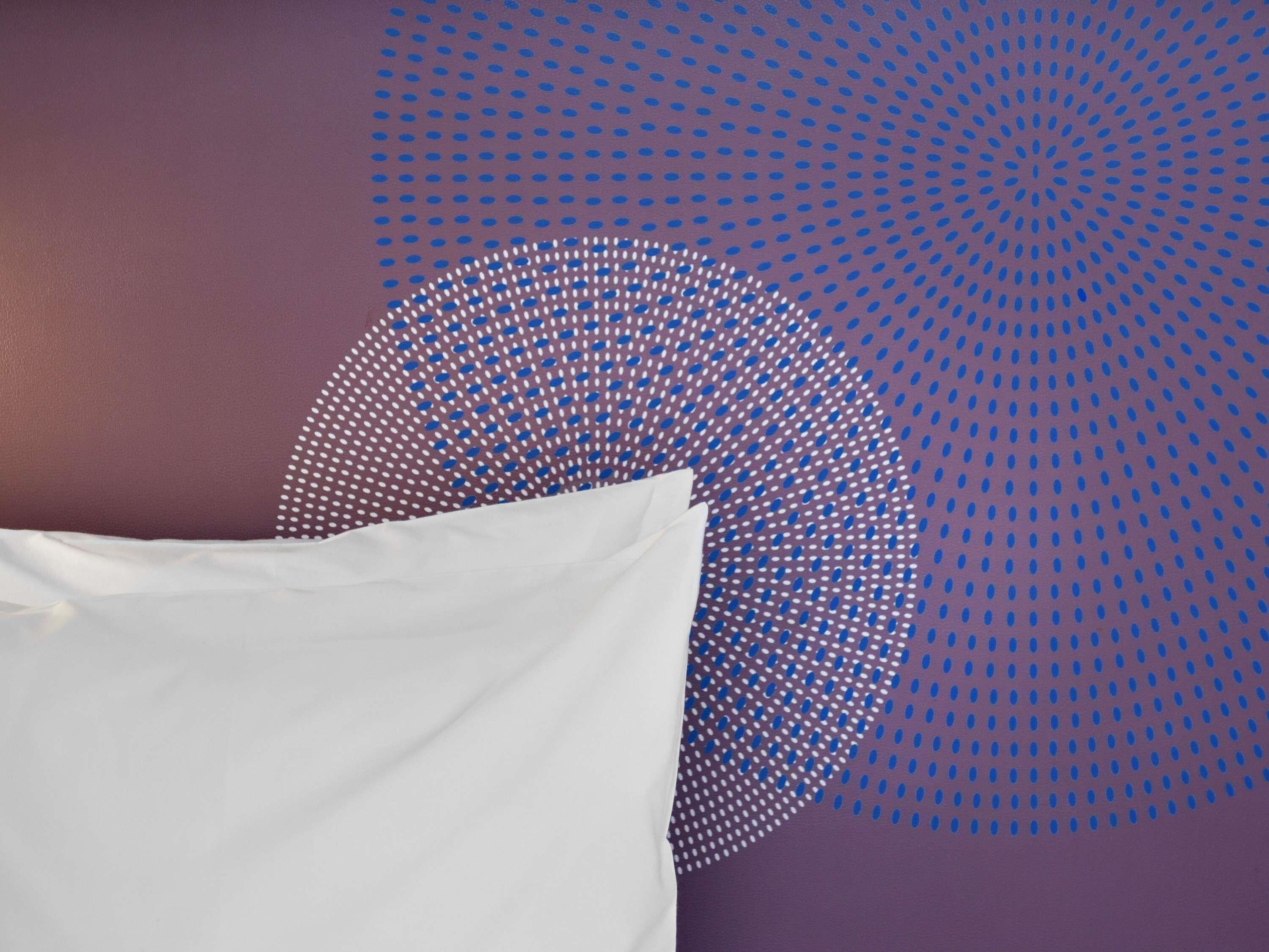 Una cama con sábanas blancas frente a una pared morada con un diseño circular de puntos azules y blancos.