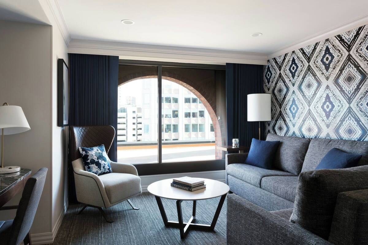 A modern hotel room with a large arched window, a grey sofa, a small round table, and a chair with a patterned cushion.