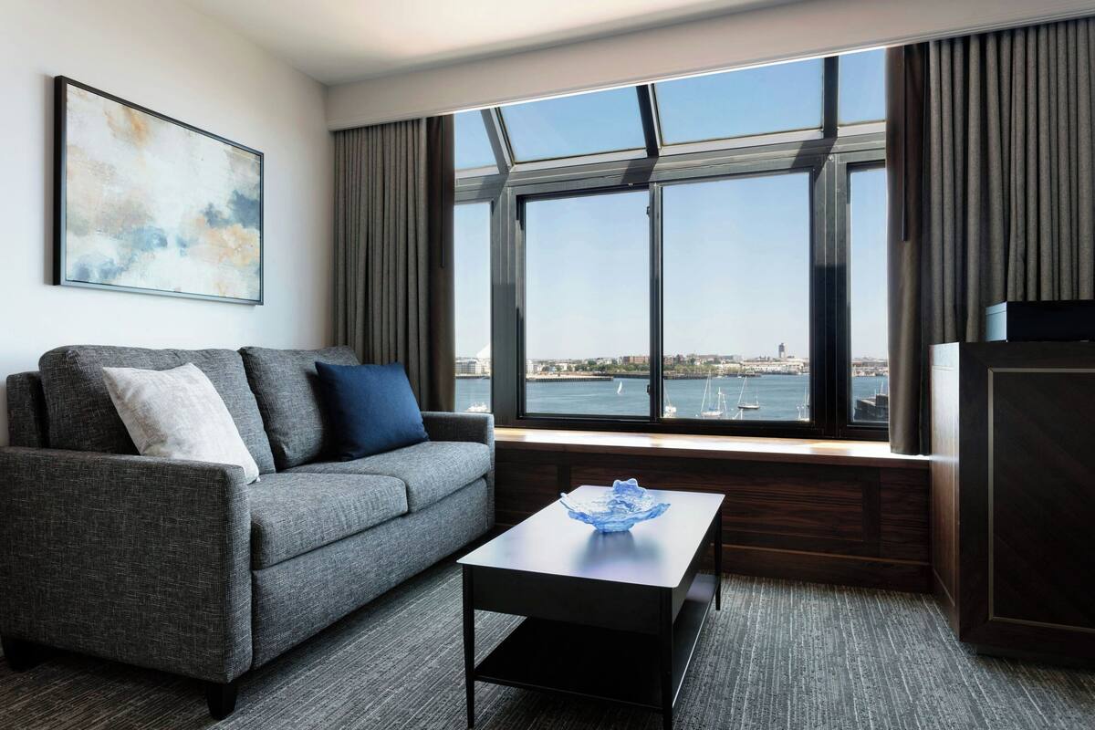 A modern hotel room with a gray sofa, a small table with a blue bowl, and a large window offering a view of a cityscape and water.