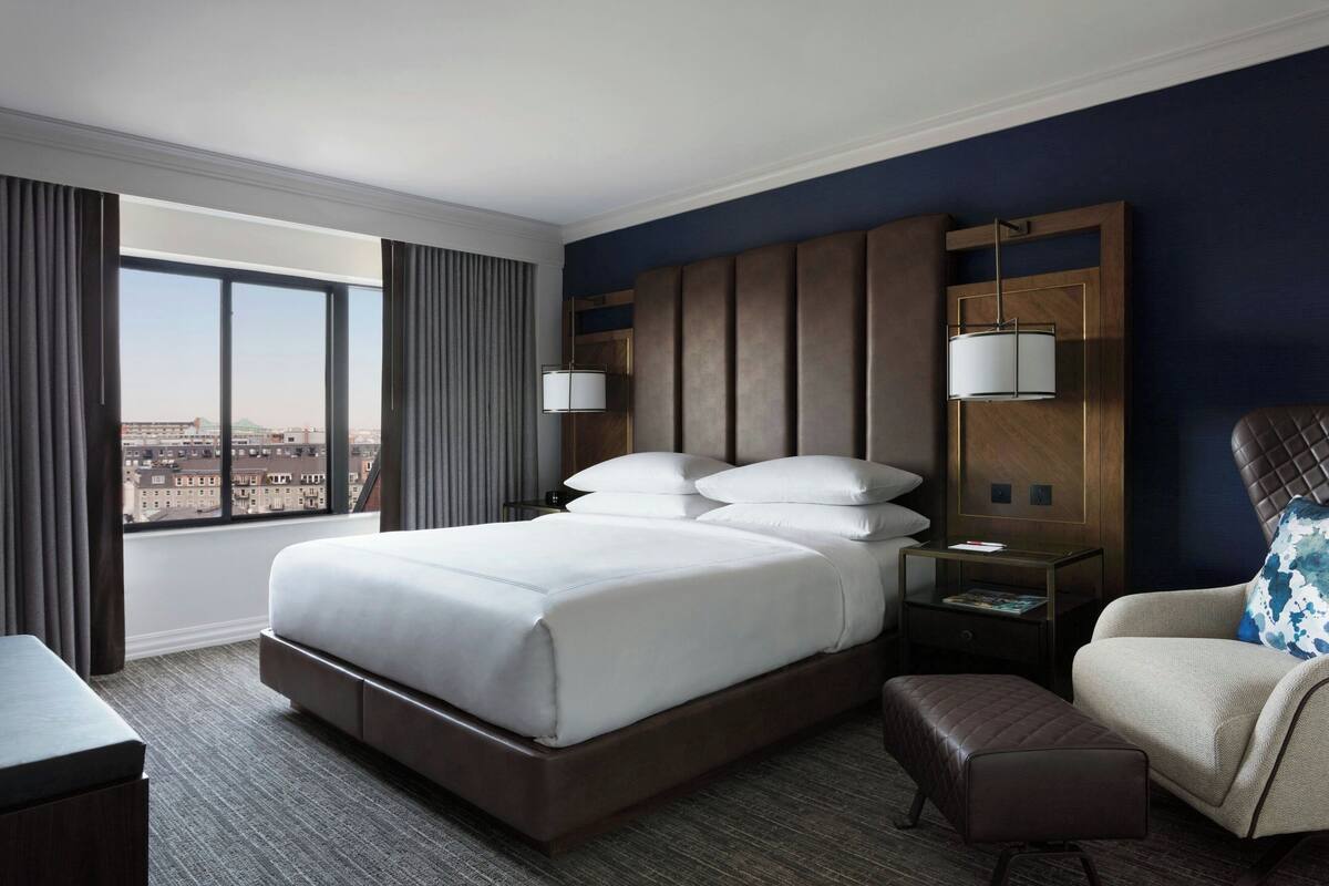 A hotel room with a large bed, a chair, a nightstand, and a view of the city.