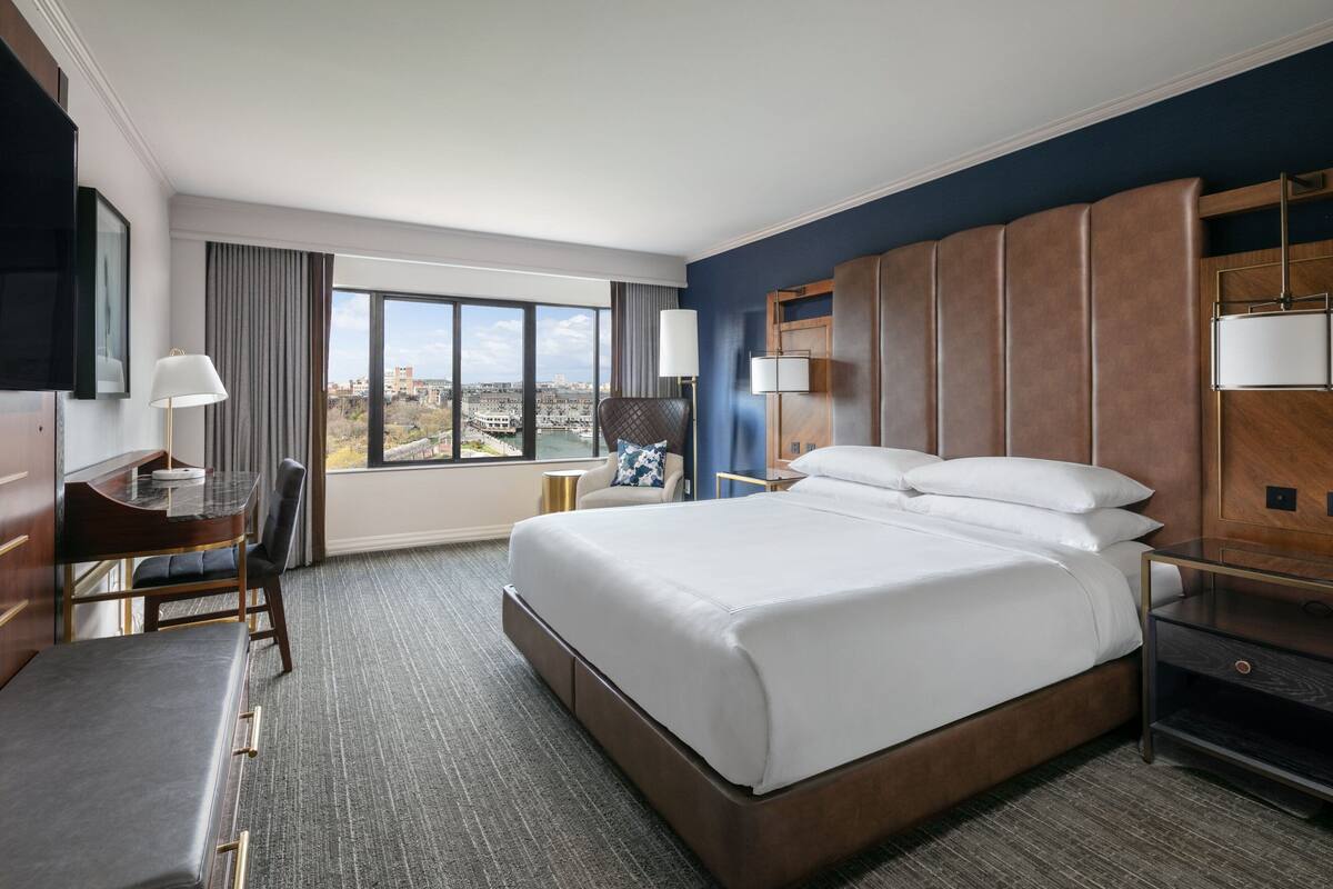 A hotel room with a large bed, a desk with a chair, a TV, and a view of the city.