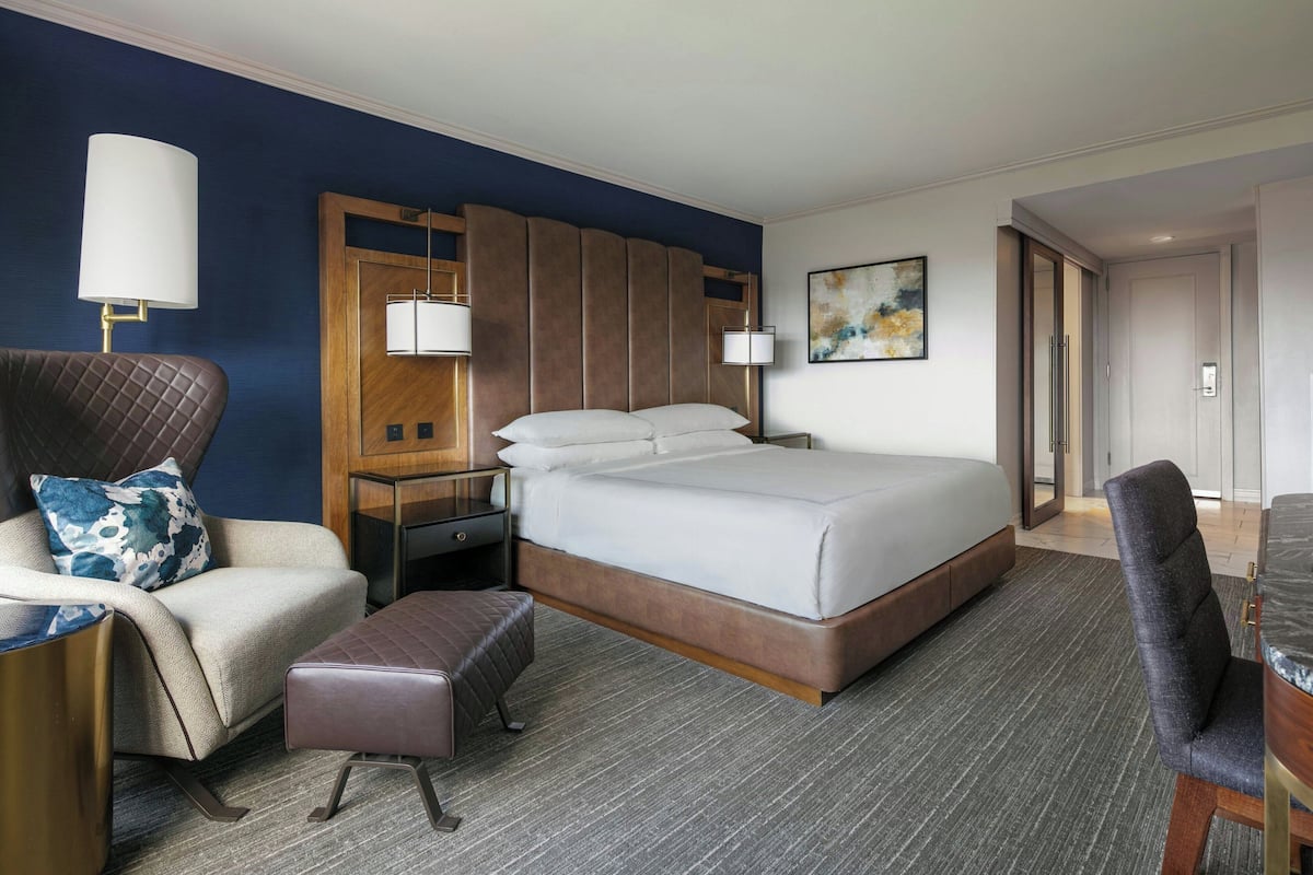 A hotel room with a large bed, a chair, a nightstand, a lamp, and a painting on the wall.