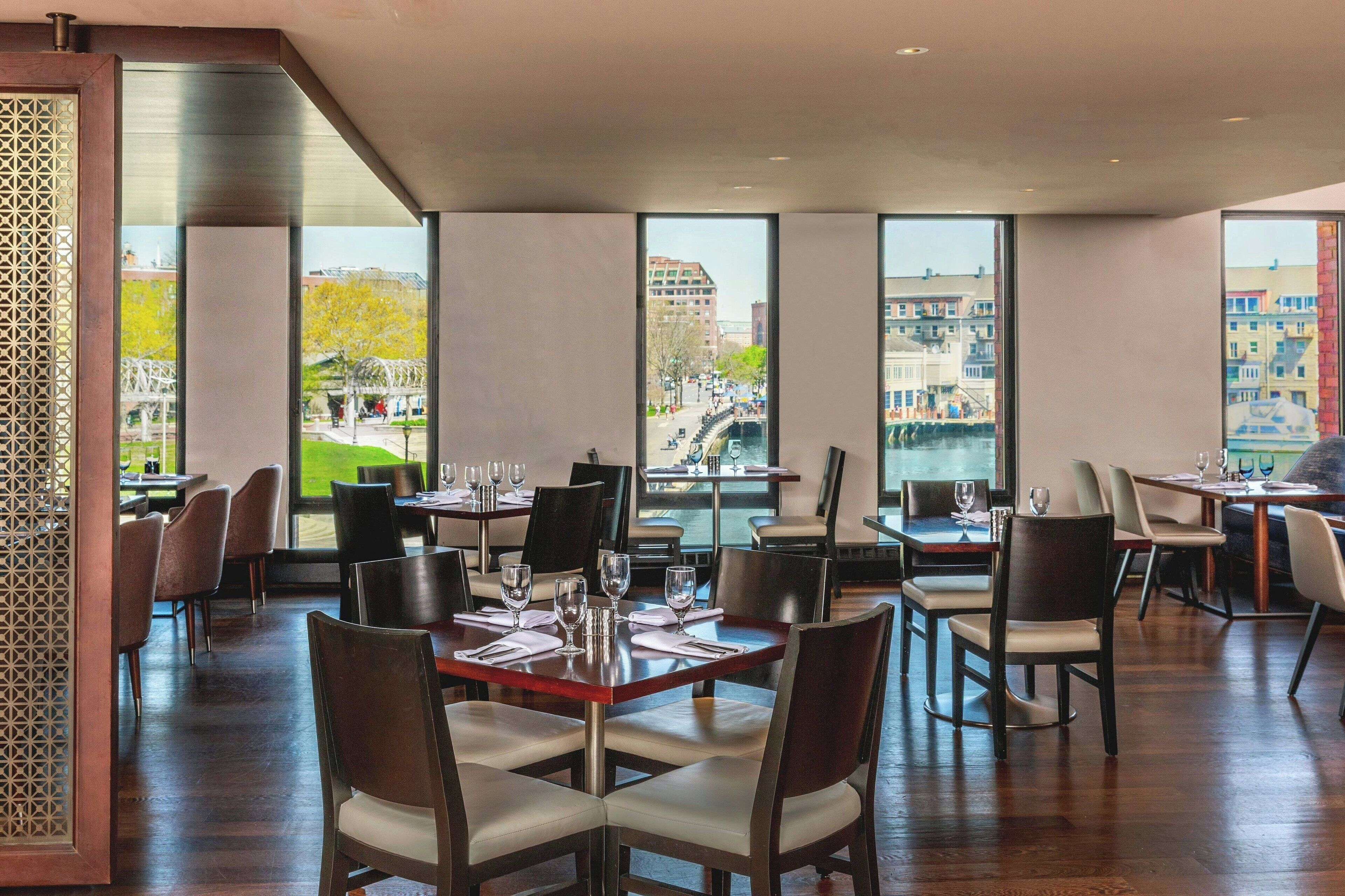 Um restaurante com mesas preparadas para refeições, grandes janelas com vista para edifícios e um parque.