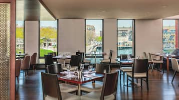 Um restaurante com mesas preparadas para refeições, grandes janelas com vista para edifícios e um parque.