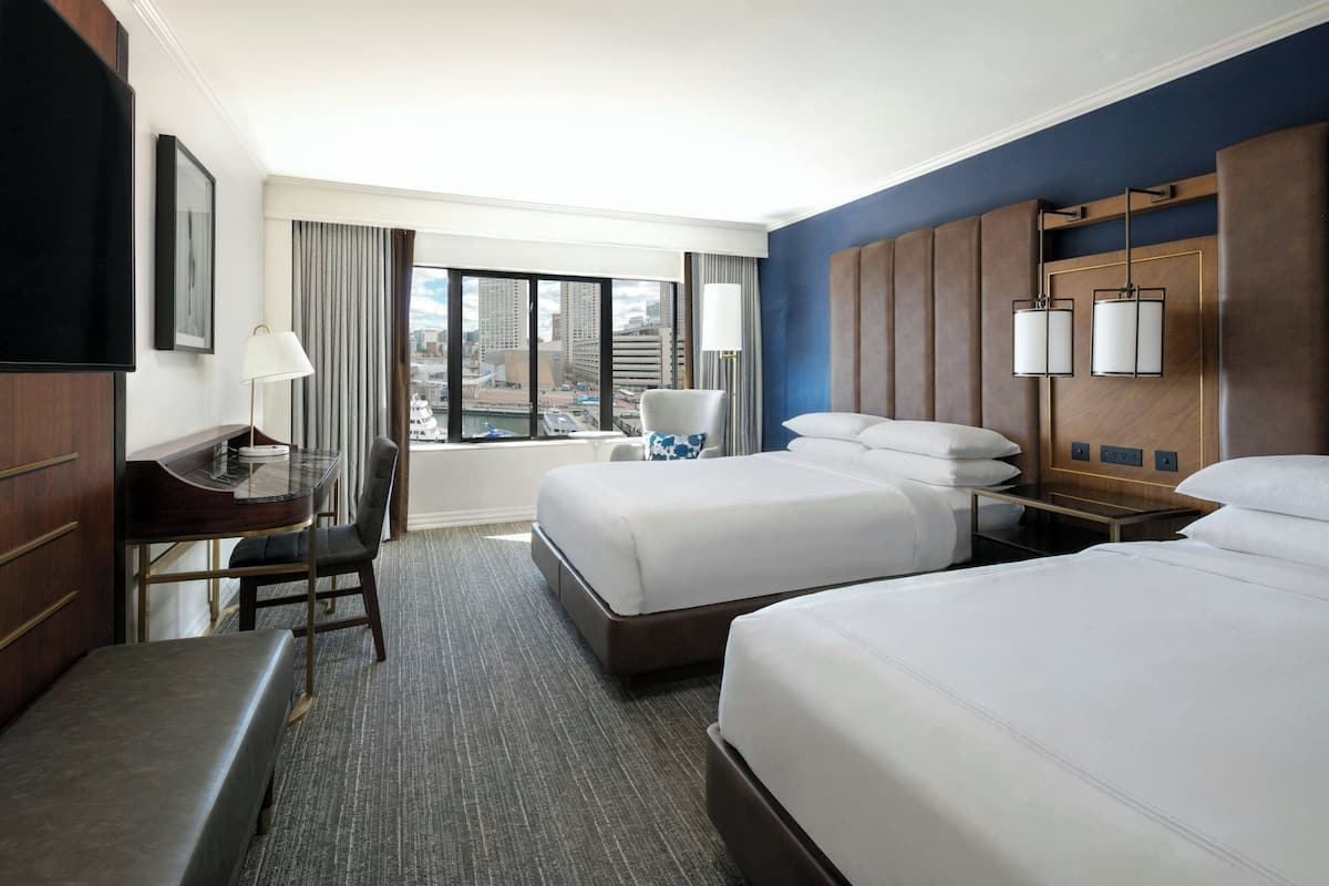 A hotel room with two beds, a desk, a chair, a TV, and a view of the city.