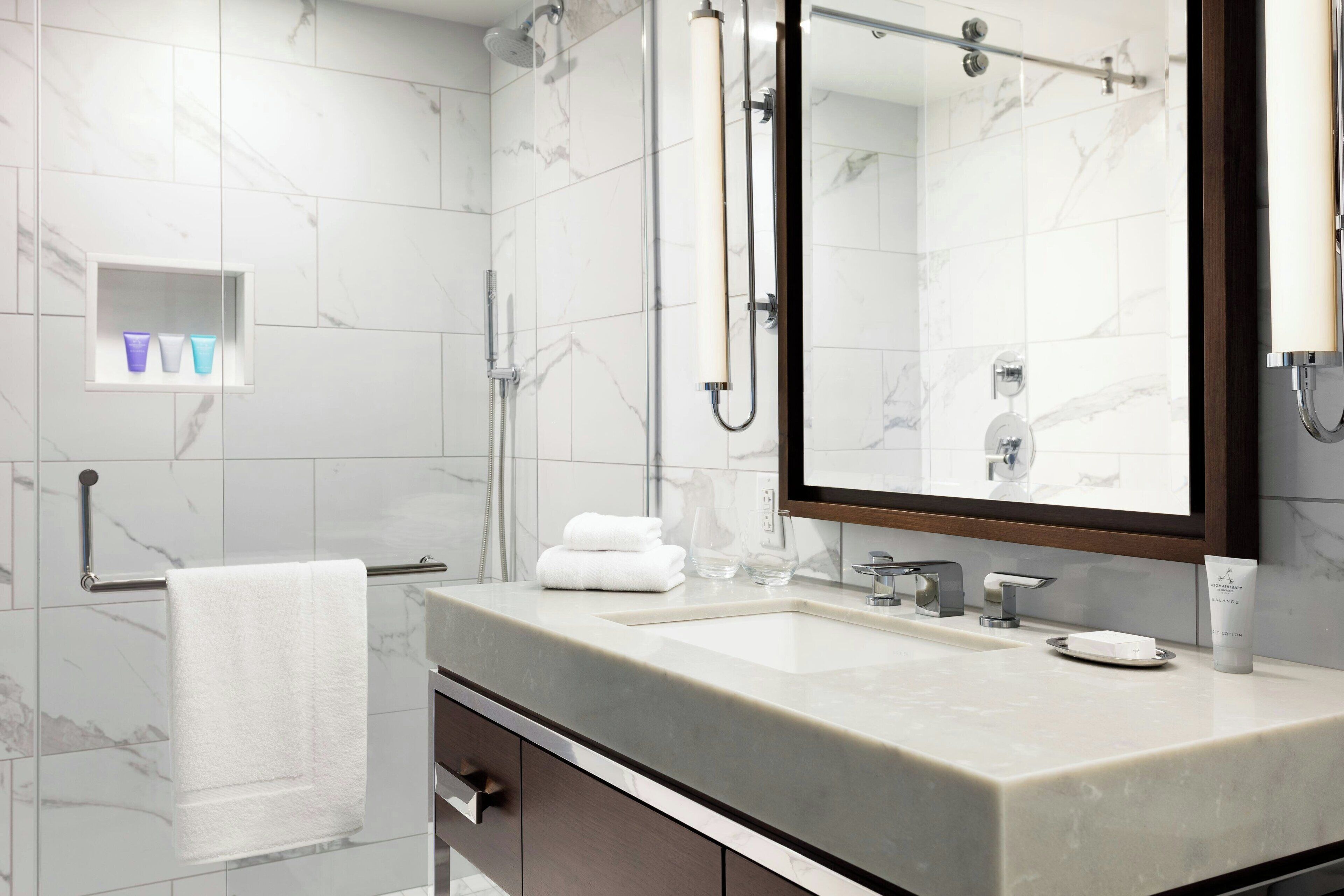 A modern bathroom with a marble-tiled wall, a large mirror, a sink with a soap dispenser, and a towel rack.