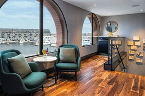 A modern room with two green armchairs, a small table with a vase, a view of a harbor, and a staircase with a mirror and wall art.