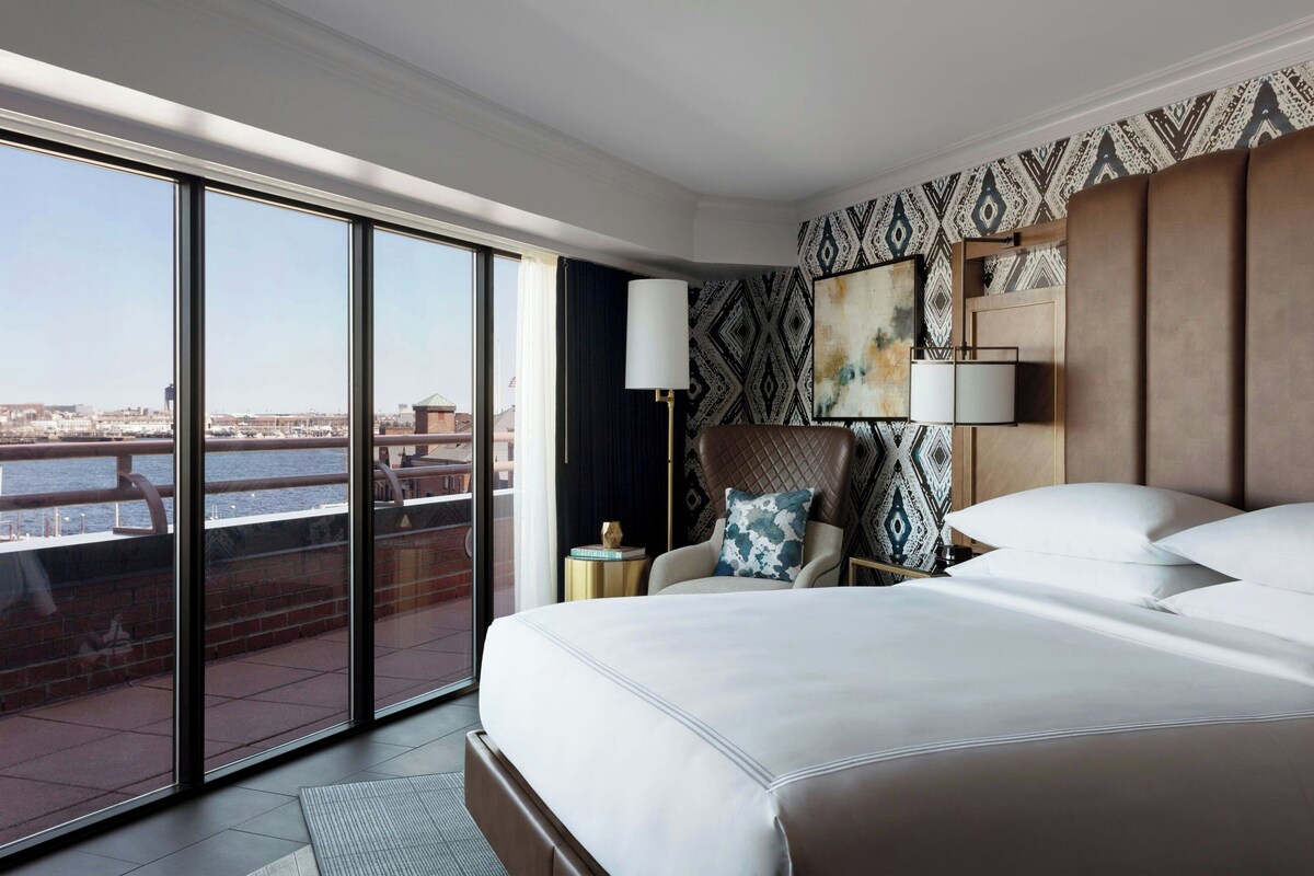 Une chambre d’hôtel avec un grand lit, une chaise, une table de chevet et une vue sur la ville, accessible par une porte coulissante.