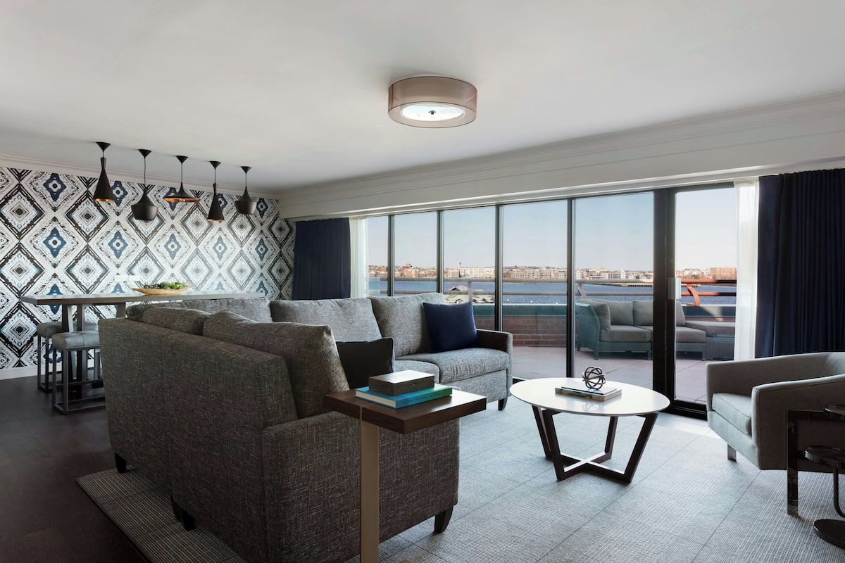 A modern living room with a sofa, armchairs, and a coffee table. There is a large window with a city view.