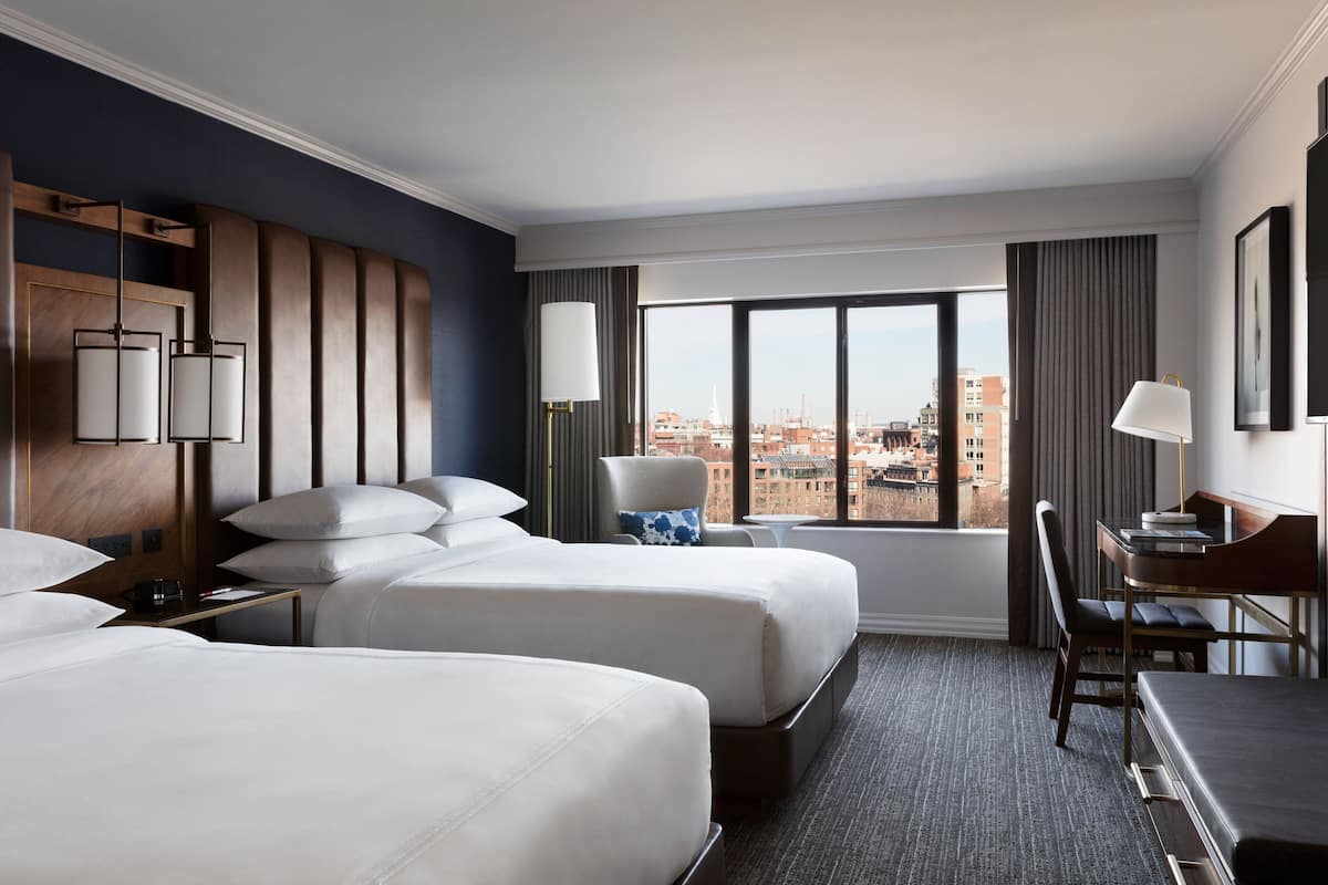 A hotel room with two beds, a desk, a chair, and a view of the city.