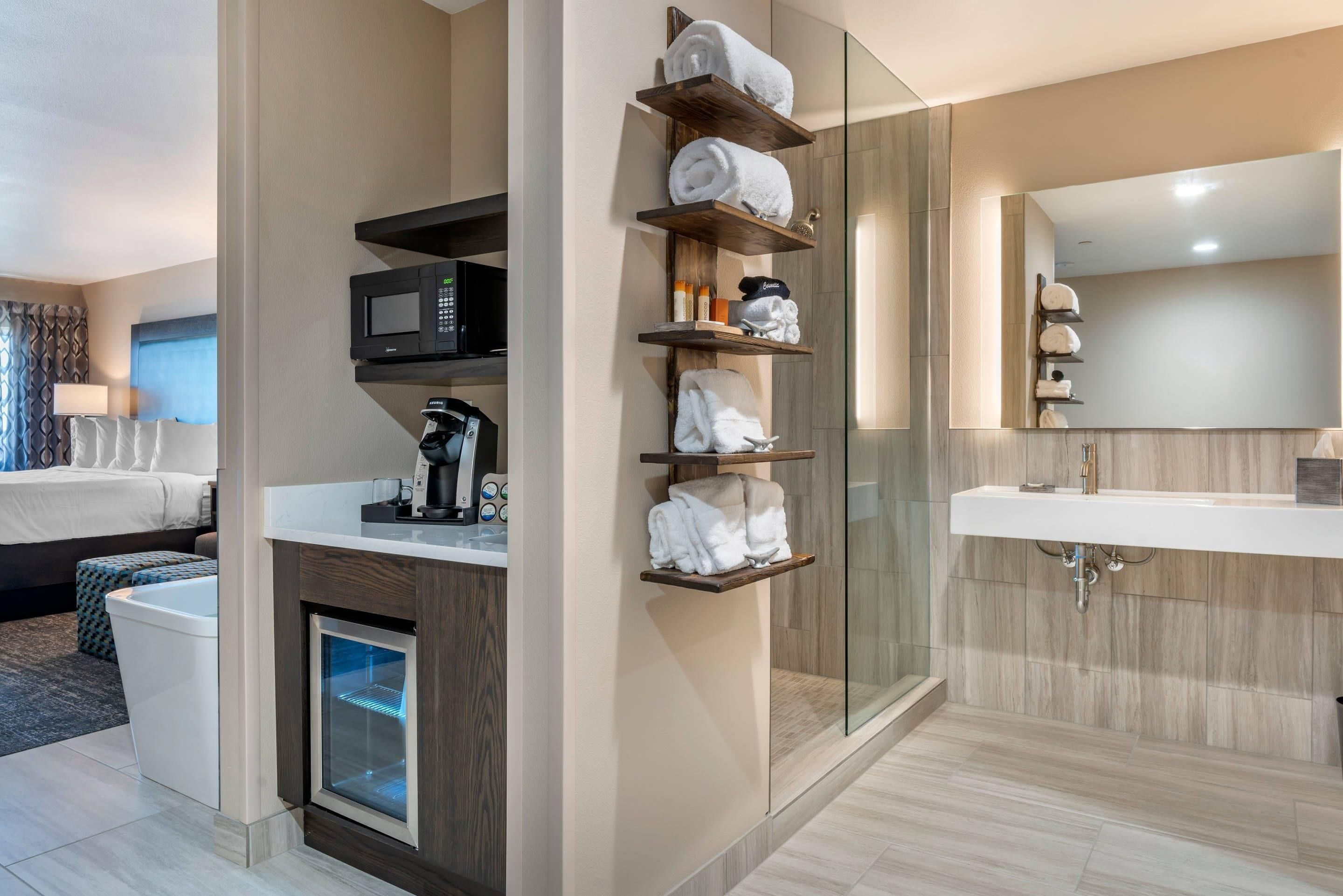 A modern hotel room with a bed, a coffee machine, a microwave, a mini-fridge, and a bathroom with a sink and glass shower.