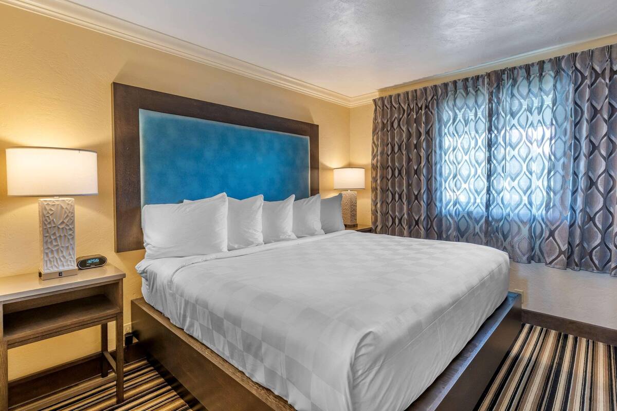 Una habitación de hotel con una cama grande, dos mesitas de noche, una lámpara de noche, un cabecero azul, cortinas estampadas y una alfombra a rayas.