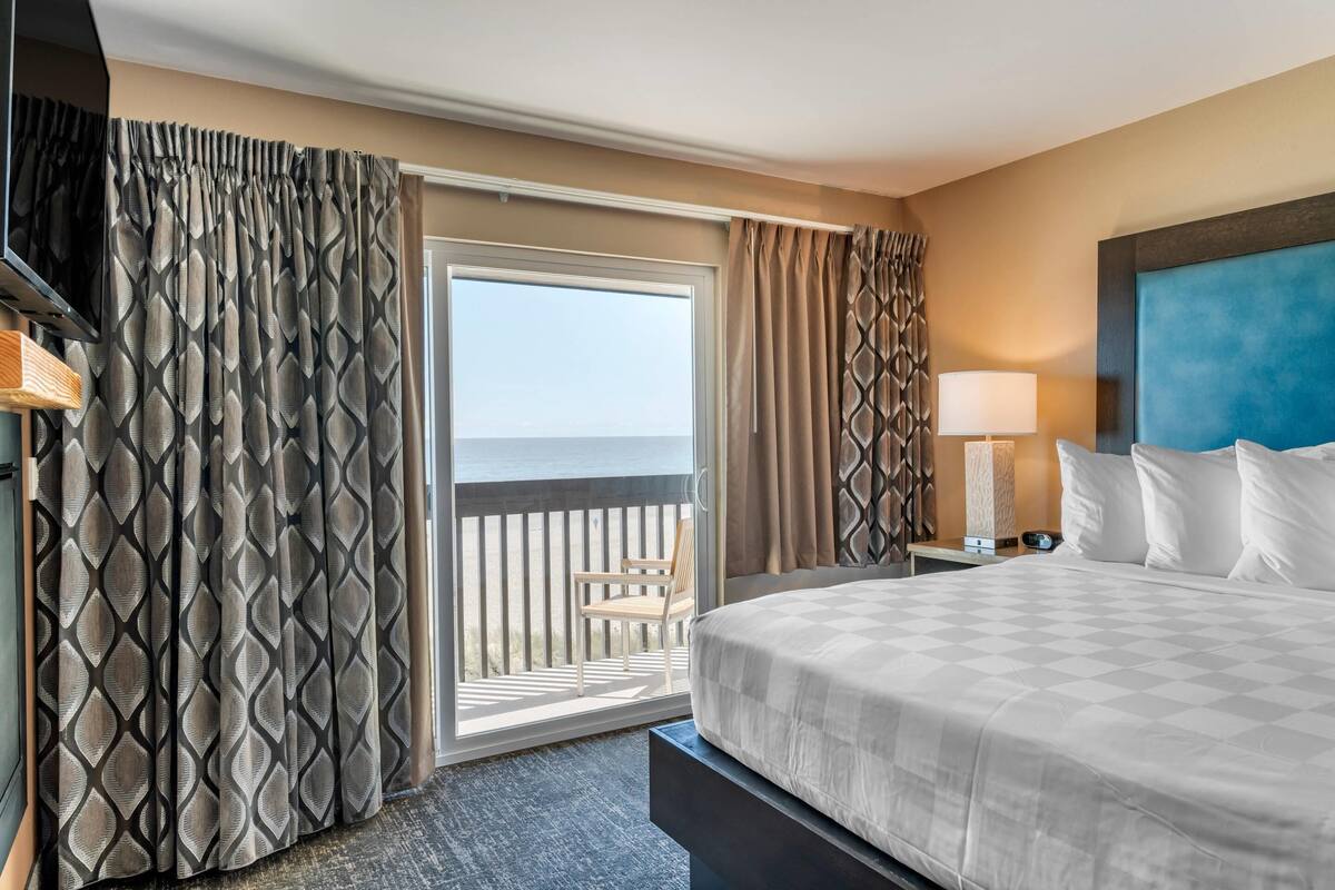 Habitación de hotel con una cama grande, una mesita de noche, una lámpara y vista al océano a través de una puerta corredera.