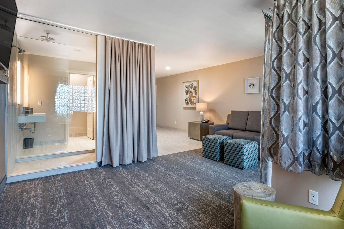 Habitación de hotel moderna con cabina de ducha de vidrio, sofá, otomana estampada y cortina decorativa.