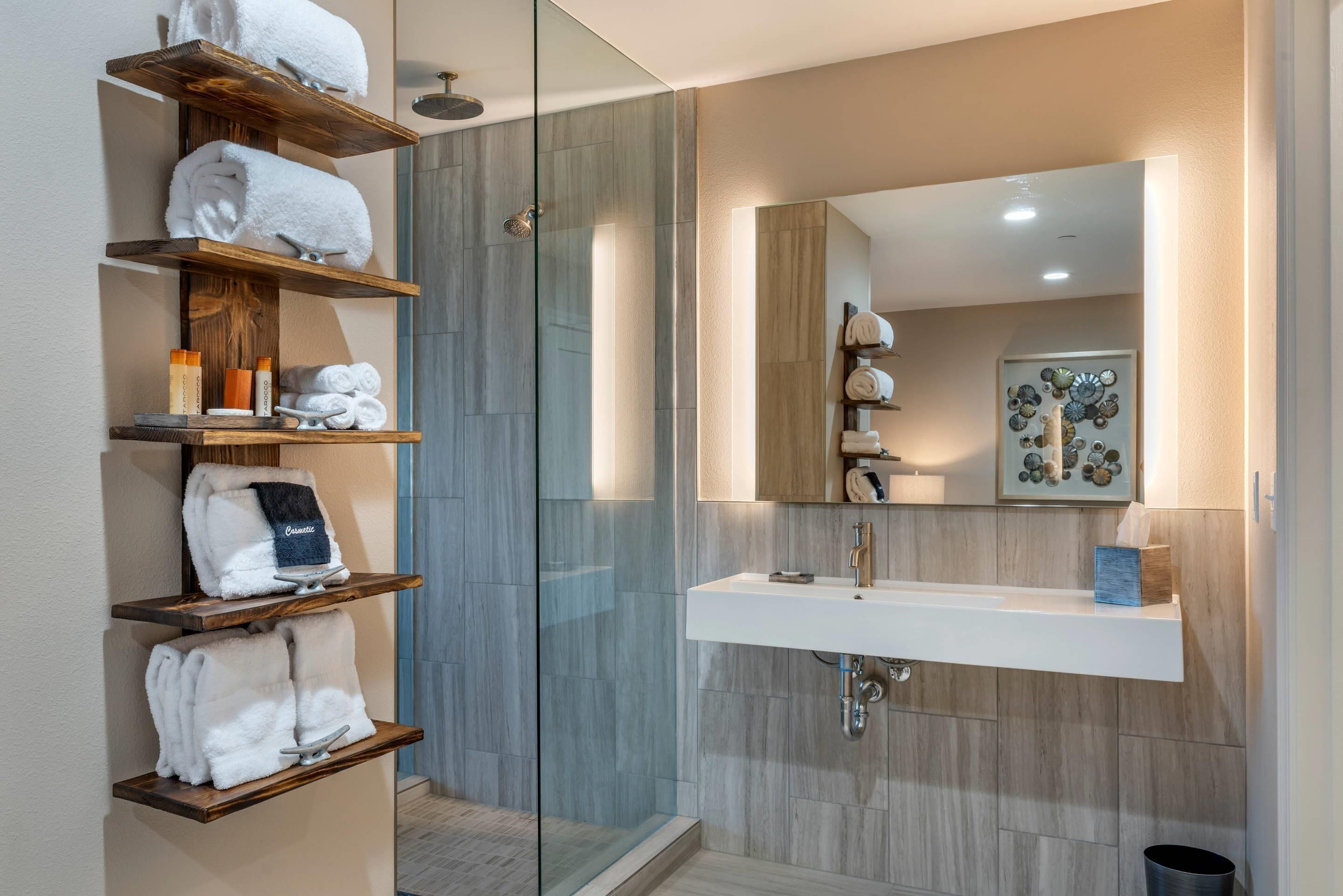 Un baño moderno con cabina de ducha de vidrio, estantes de madera con toallas, un lavabo blanco y un espejo con un adorno encima.