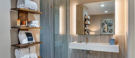 A modern bathroom with a glass shower enclosure, wooden shelves with towels, a white sink, and a mirror with a decorative piece above it.