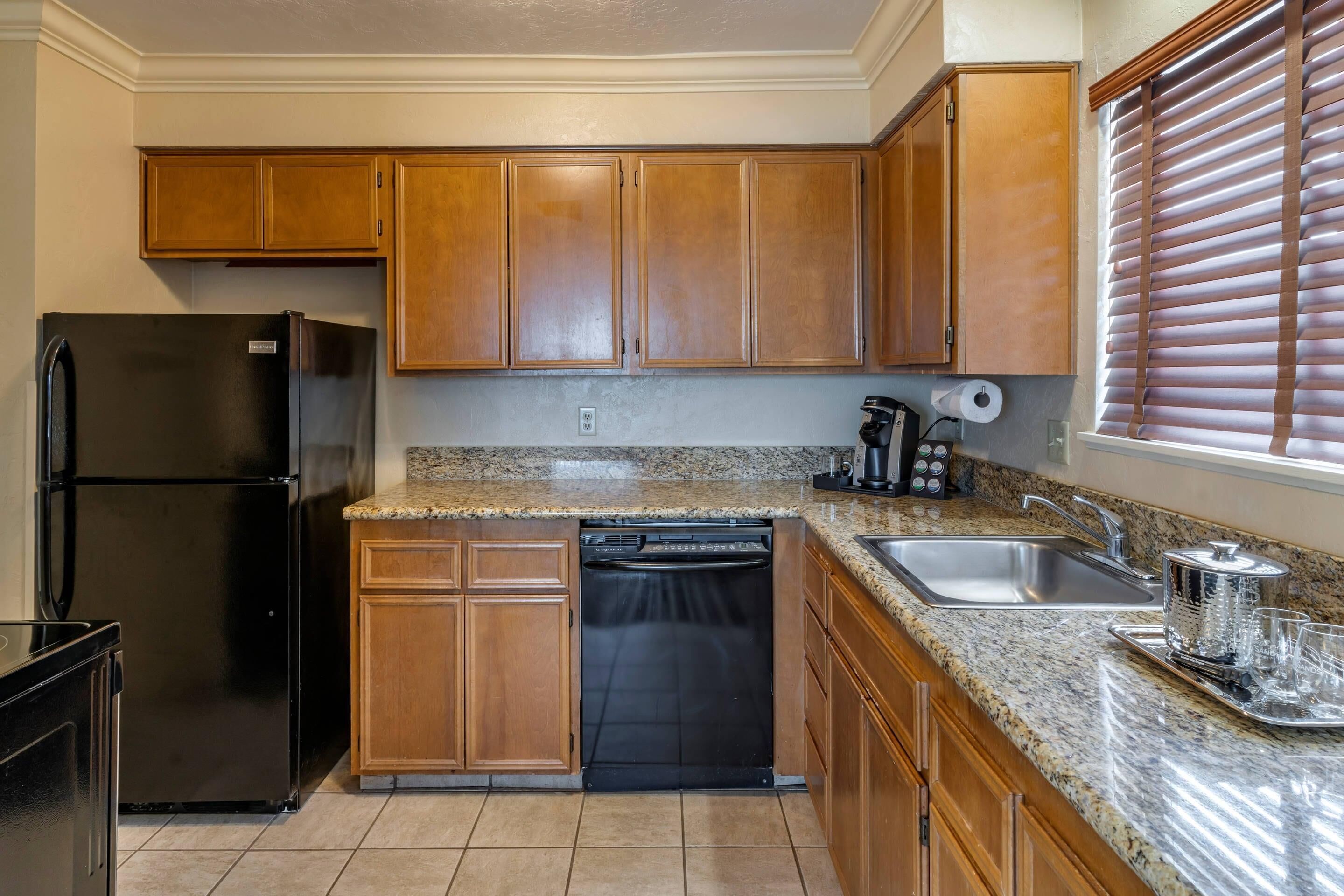 A kitchen with wooden cabinets, a black refrigerator, a dishwasher, a granite countertop, a sink, and a window with blinds.