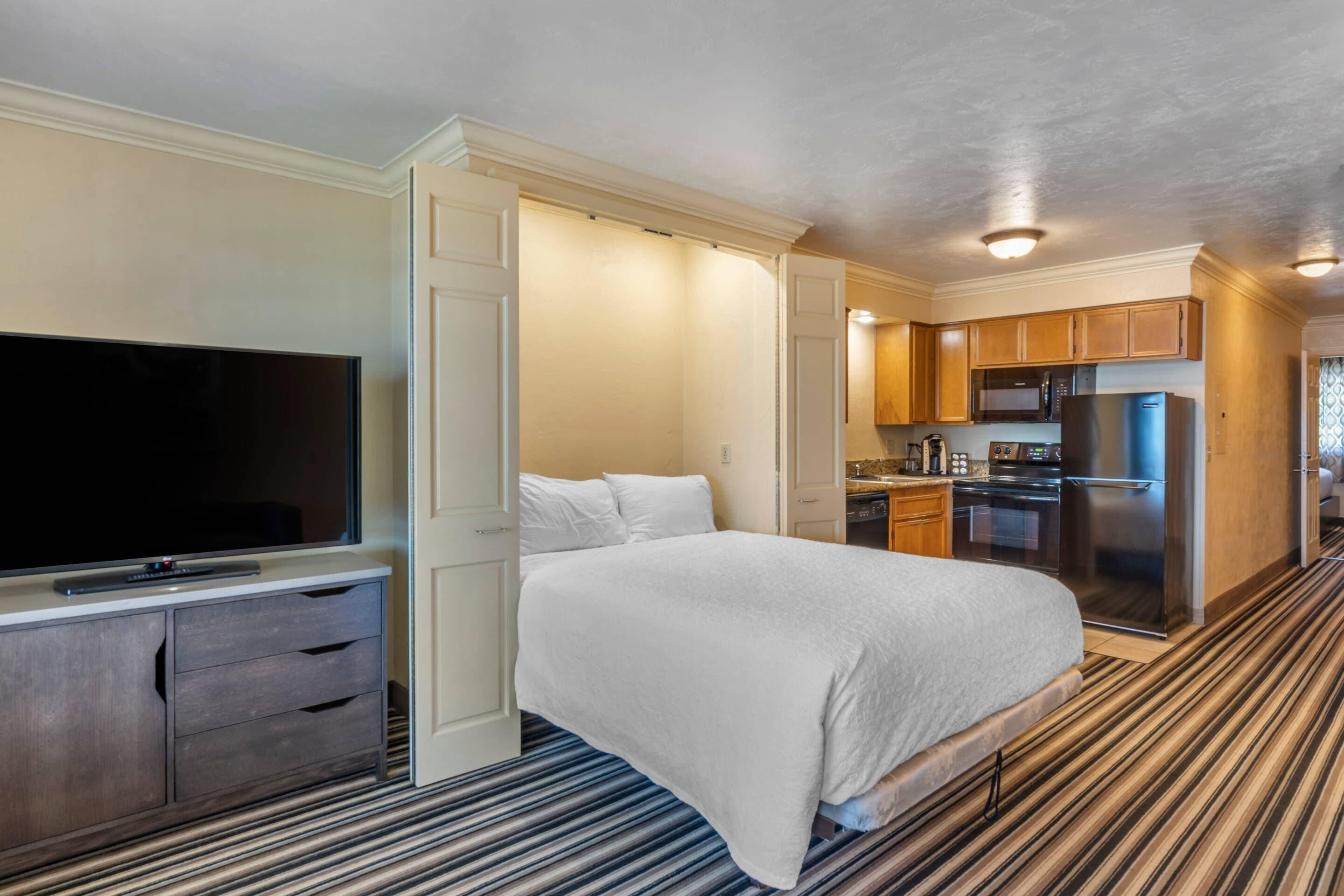 A hotel room with a bed, a TV, a kitchenette, and a striped carpet.