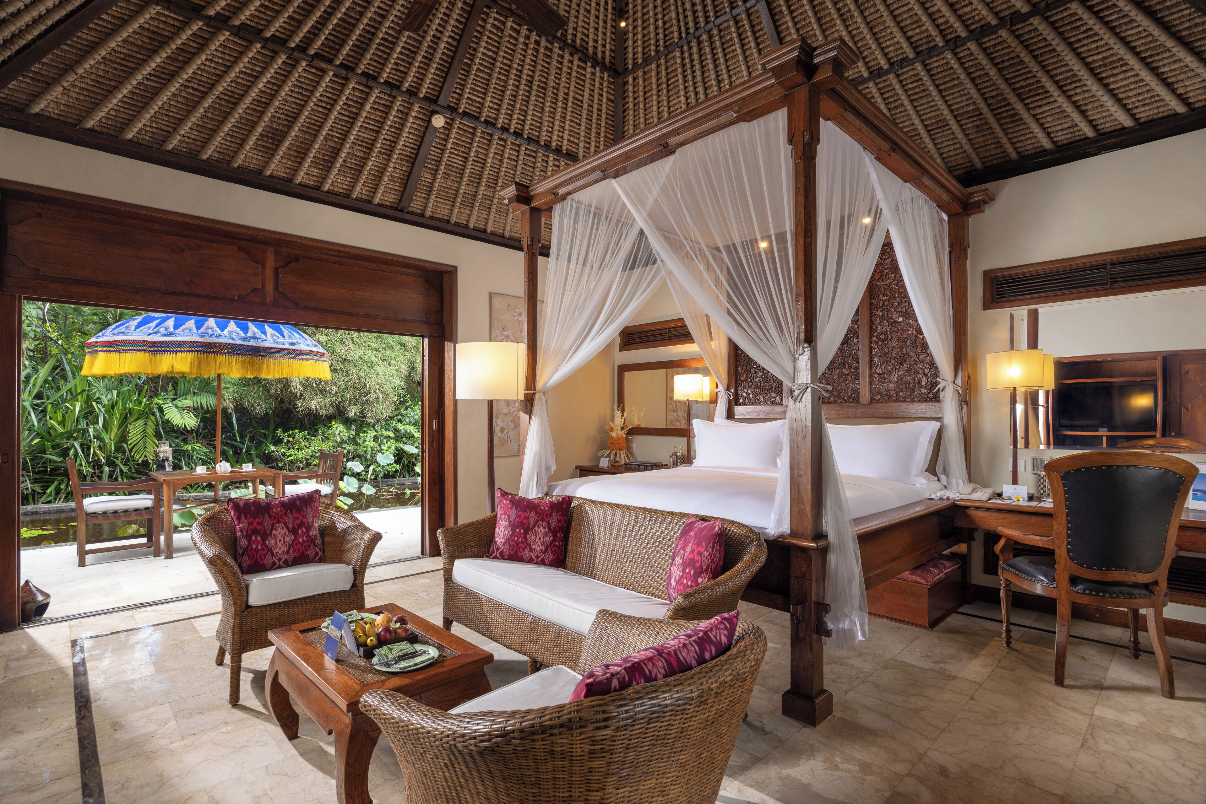 A four-poster bed with a canopy, a sitting area with wicker furniture, a desk with a chair, and a view of greenery outside.