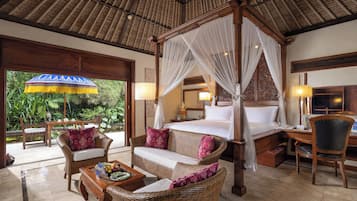 A four-poster bed with a canopy, a sitting area with wicker furniture, a desk with a chair, and a view of greenery outside.